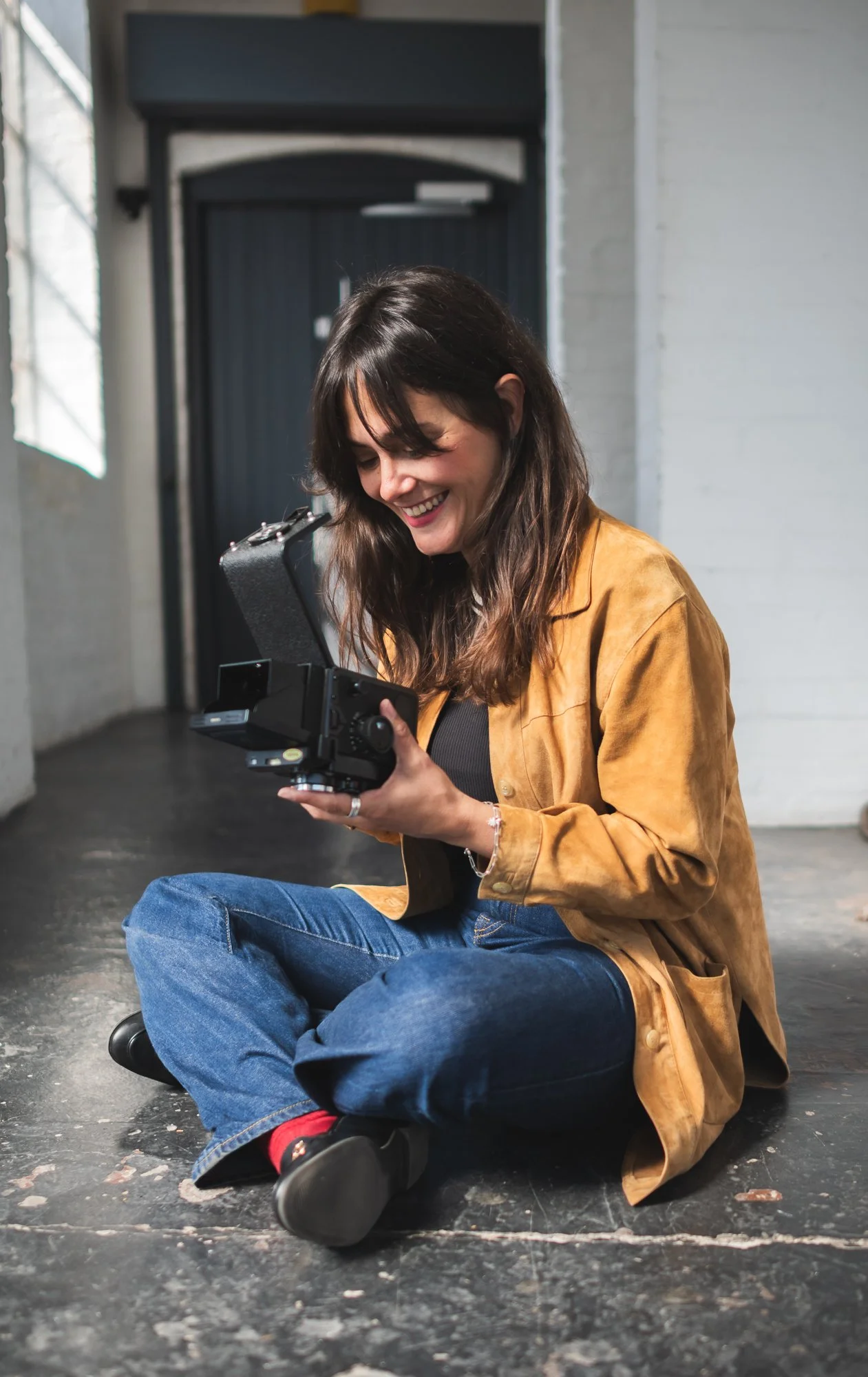A woman with brown hair, wearing a tan jacket, blue jeans, and black shoes with red socks, sat cross-legged on a black floor, looking down at a camera with a smile.