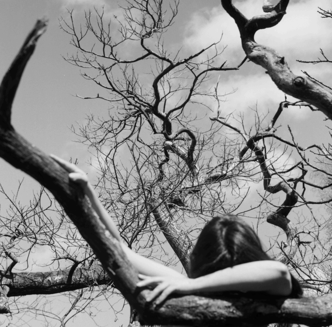 A person with long hair leaning on a tree branch, with their arm stretched out holding another branch, against a cloudy sky in black and white.