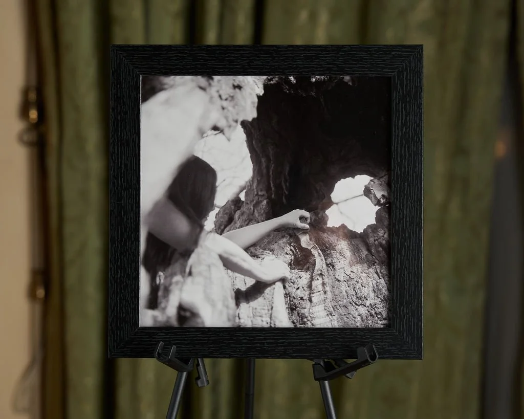 A black-and-white photograph of a woman with long hair climbing a rocky surface, holding onto a handhold, with a large rock formation and an opening in the rock in the background.