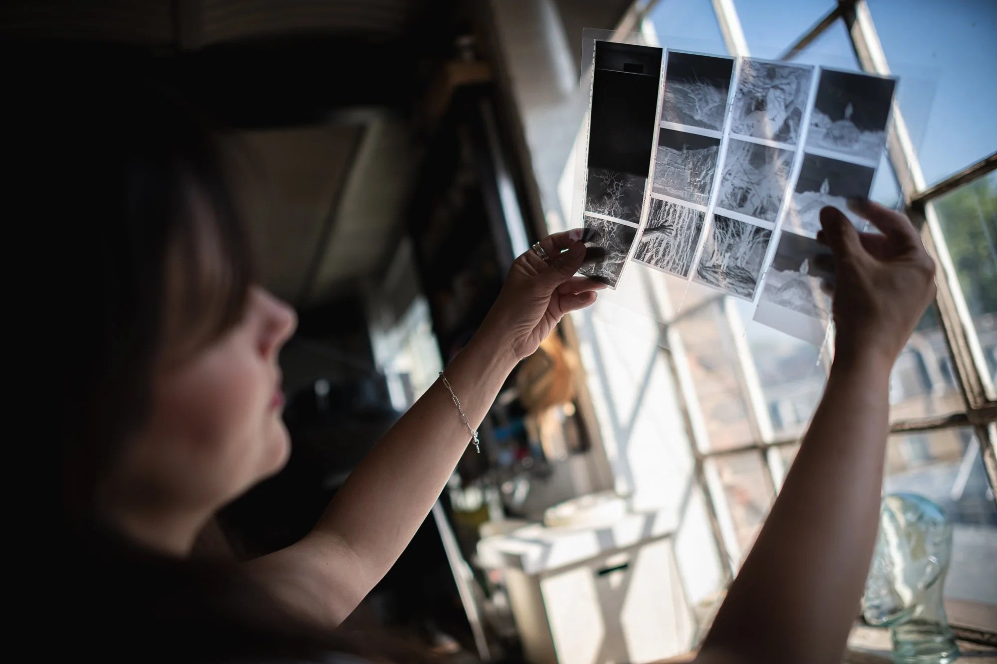 A woman holds black-and-white photographs of trees and landscapes inside a house near a window.
