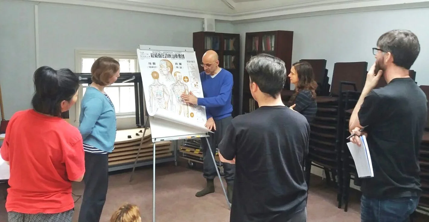 Photograph of Nik Kyriacou stood teaching using a Shiatsu meridian map with three students watching and listening