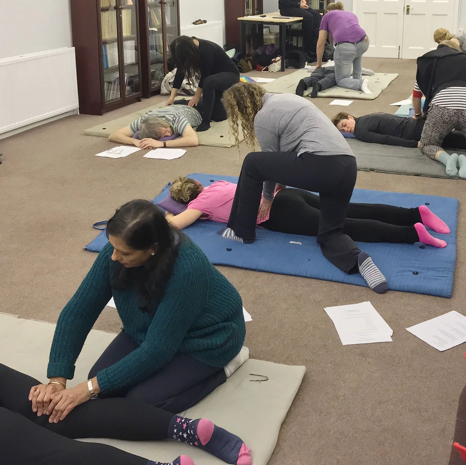 Five pairs of Shiatsu students practicing with practitioner kneeling whilst applying finger pressure to lower back of partner who lies face down on futon