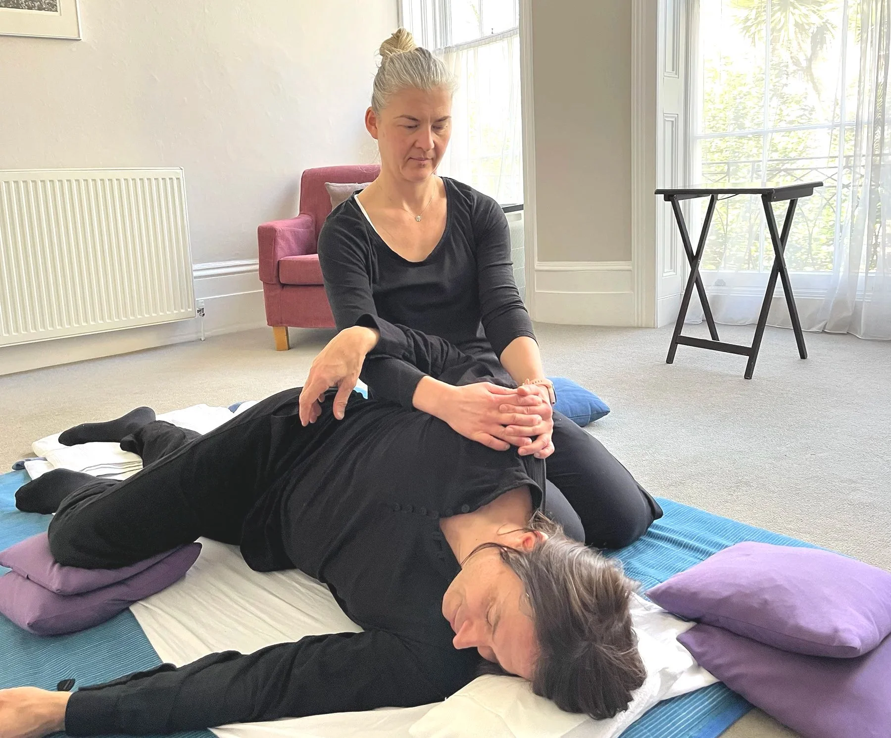 Photograph of a Shiatsu session in a bright airy treatment room, the practitioner stretching the client's shoulder who is lay on their side