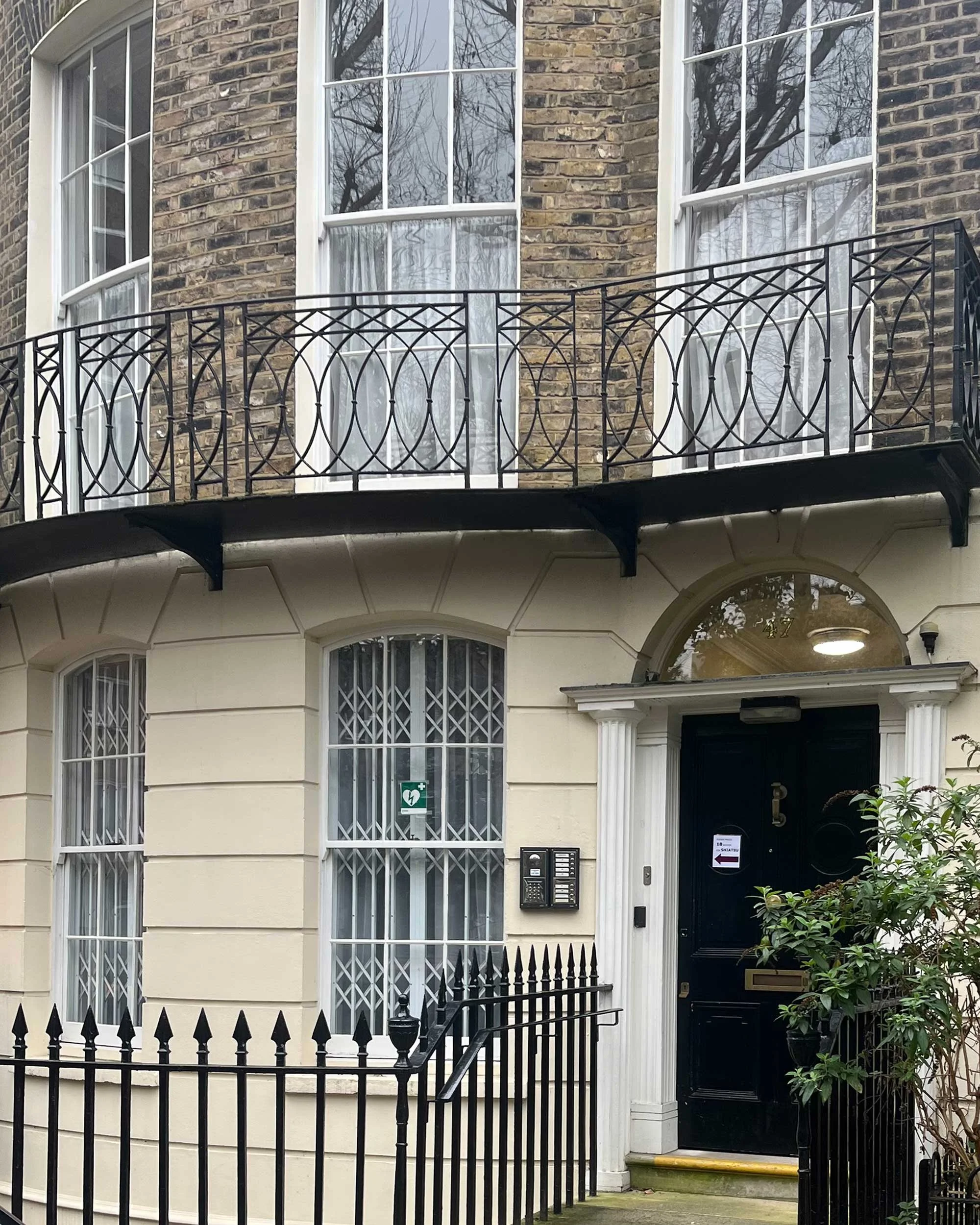 Photograph showing exterior of a historical Georgian building featuring tall paned sash windows, ornate black metal railings, cream panelling and two ornate white columns on either side of a traditional painted black door