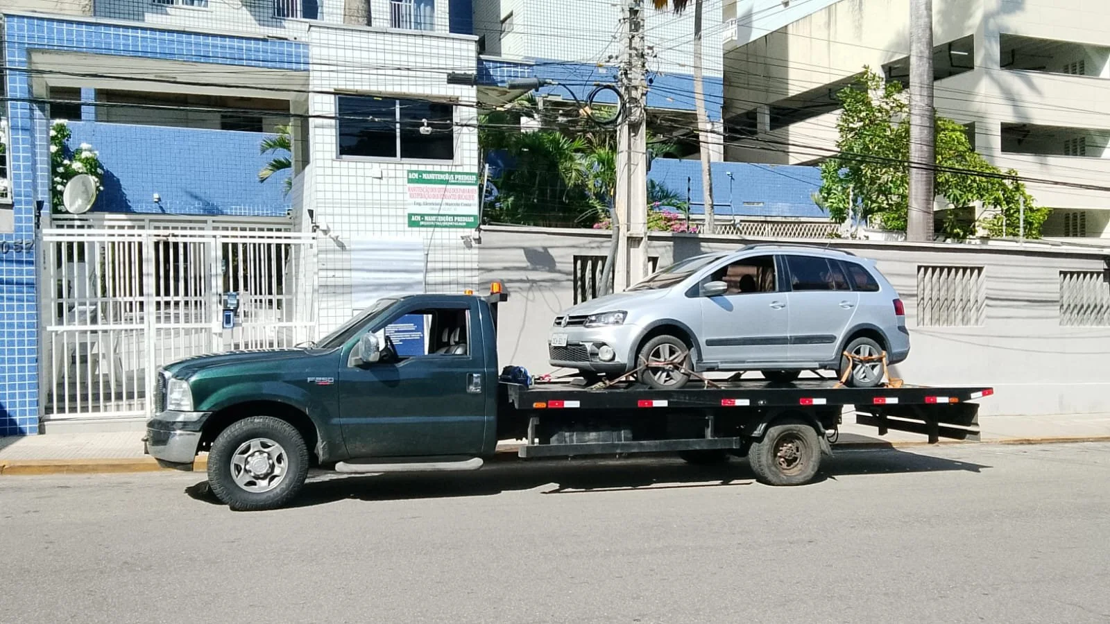 Carro prata sendo transportado por um caminhão na rua, com prédios e árvores ao fundo.