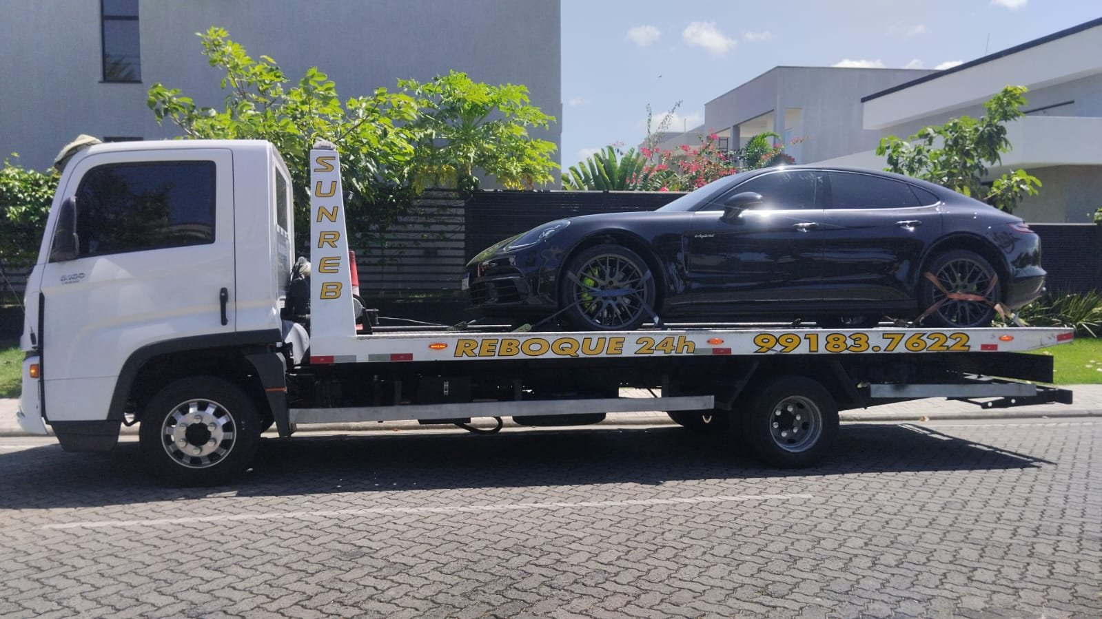 Carrinho de transporte de veículos com um carro preto em cima, estacionado na rua pública, com vegetação e edifícios modernos ao fundo.