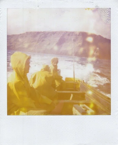 Three people in yellow raincoats are on a boat off the coast of Easter Island, with a body of water and hills in the background, during daylight.