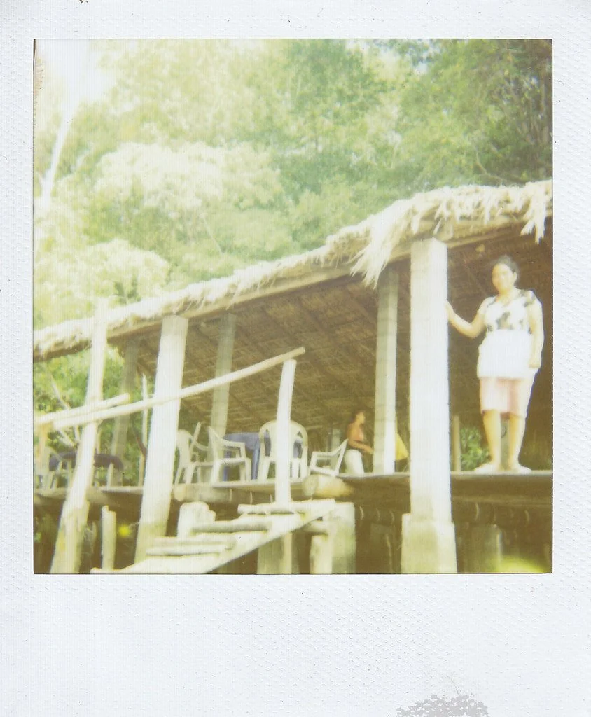 A woman standing outside a thatched-roof wooden hut on stilts, with trees and greenery in the background.