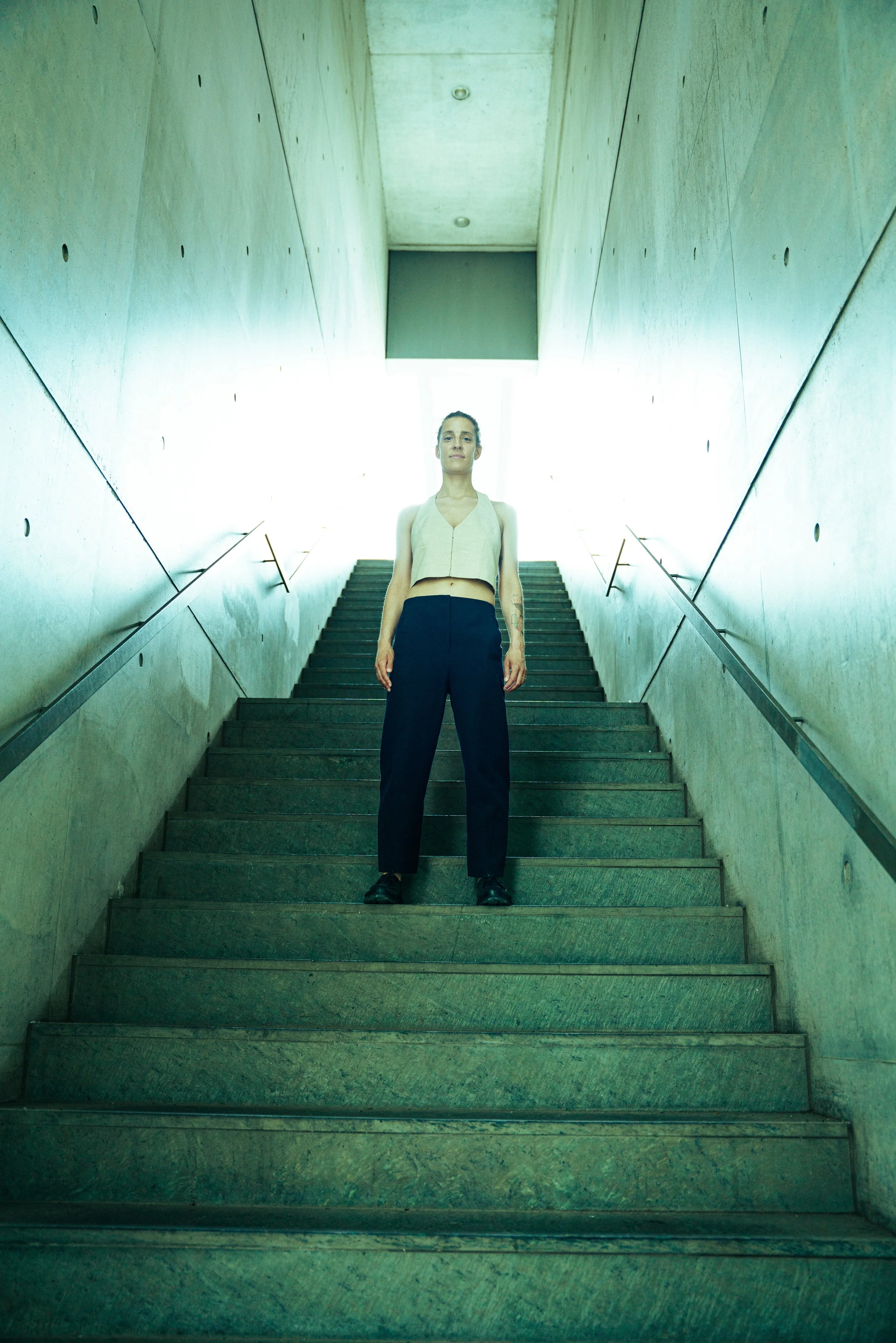 A woman standing on a wide staircase with concrete walls on either side and a bright light at the top, wearing black pants and a tan sleeveless top.