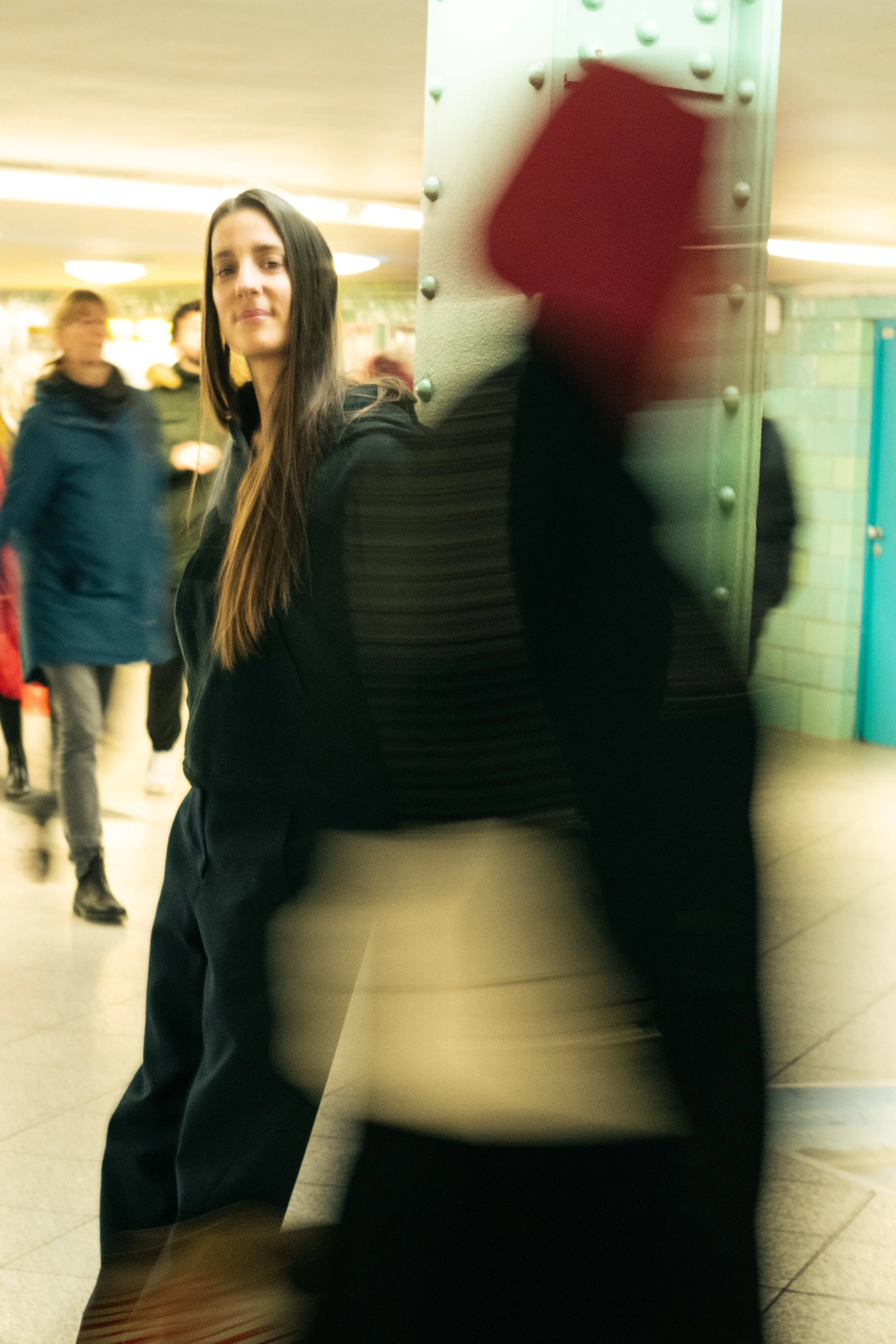 A young woman with long brown hair standing by a large pillar in a busy indoor public space, with people walking by and a green tiled wall in the background.