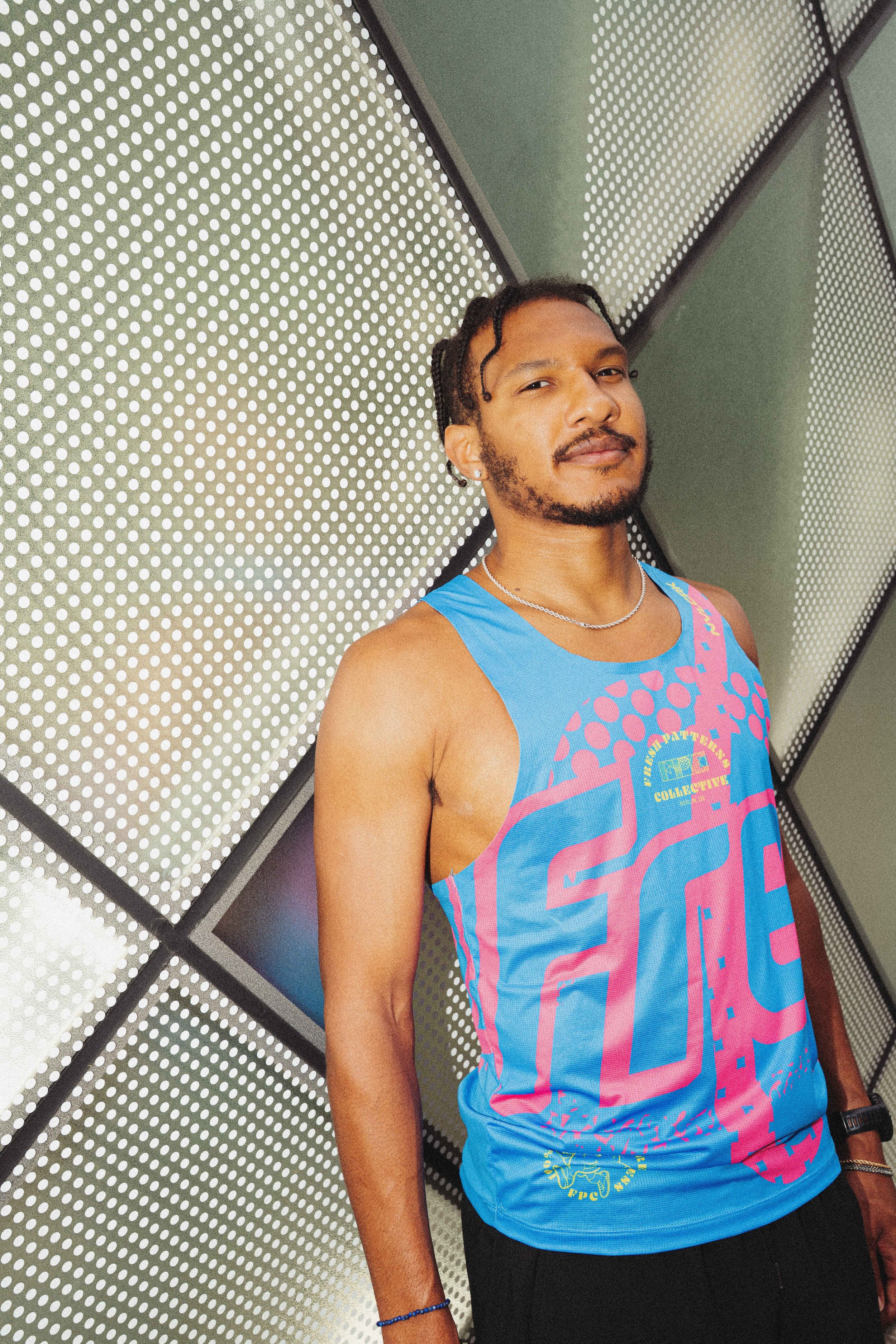 A person wearing a colorful blue tank top with pink and yellow designs, standing in front of a perforated metal wall with circular holes and a geometric pattern.