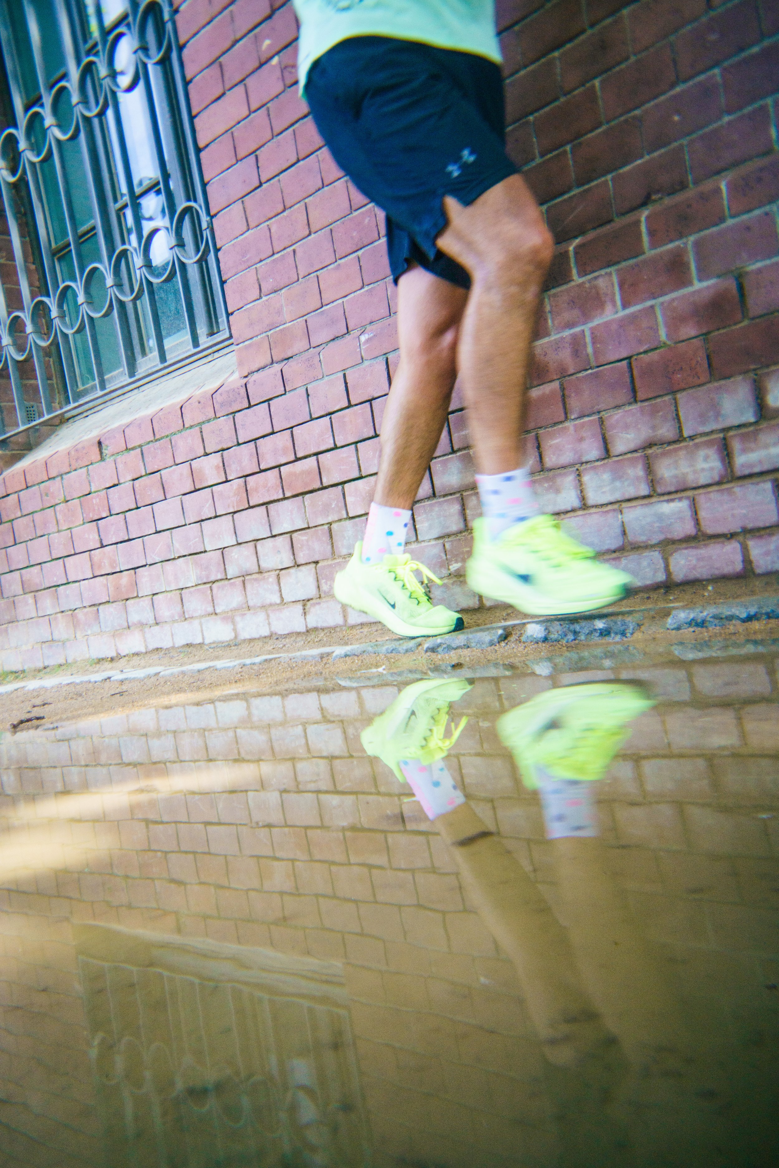 Person standing next to a brick wall, wearing neon yellow running shoes, black shorts, a light yellow shirt, and polka dot socks, with their reflection visible in a puddle on the ground.