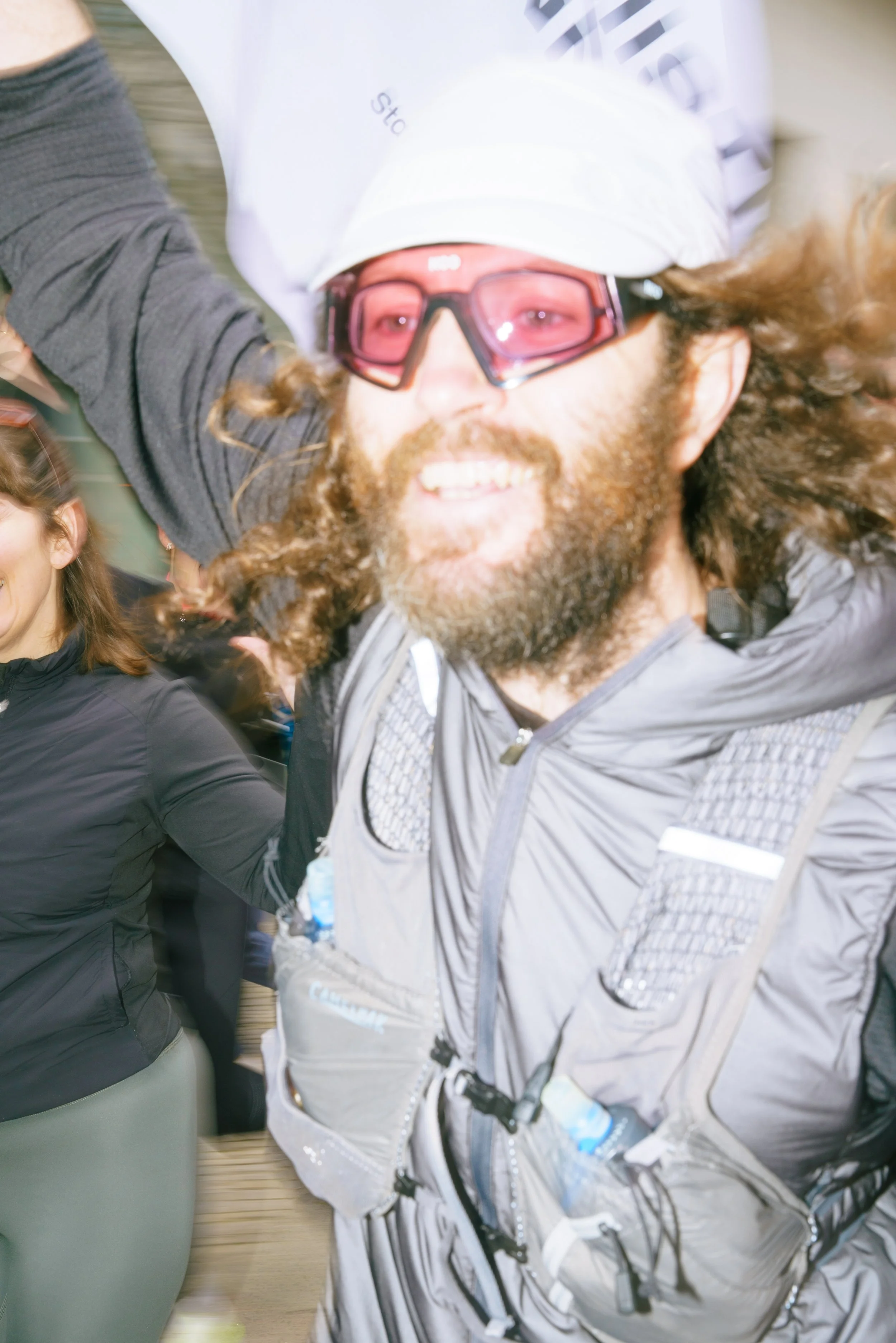 Close-up of smiling man with long hair, beard, wearing sunglasses, white cap, and gray running vest.
