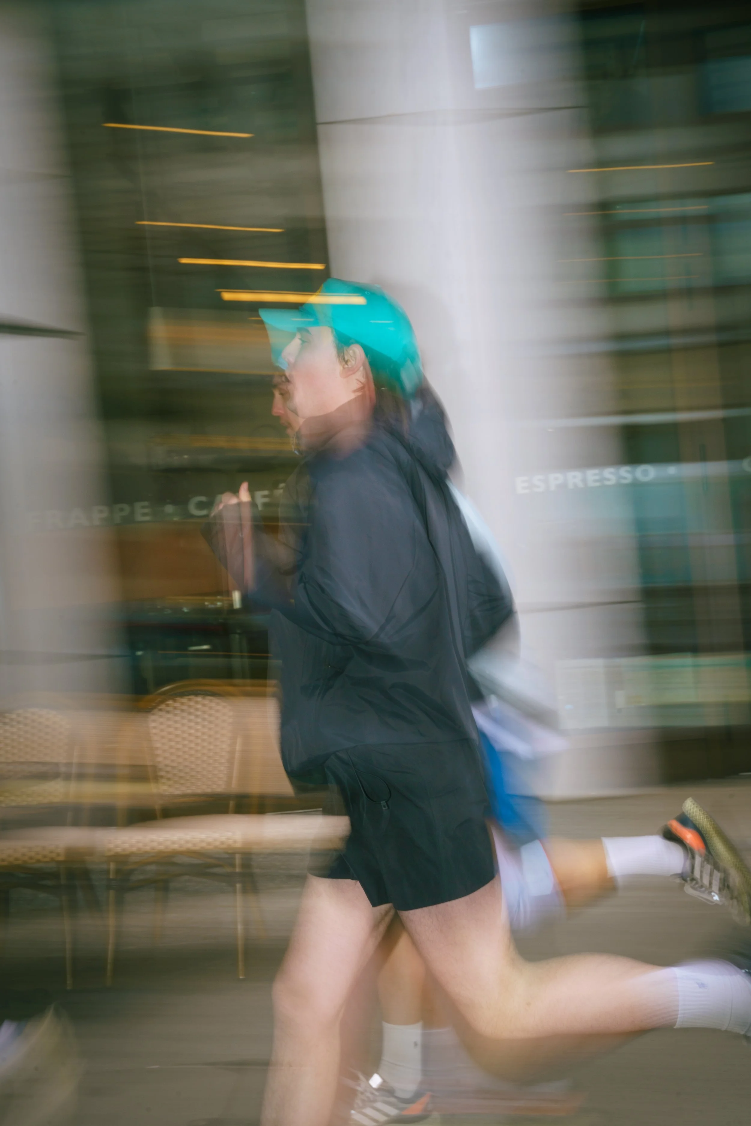 A man running outside a cafe wearing a blue helmet, black jacket, shorts, and white socks, with motion blur indicating fast movement.
