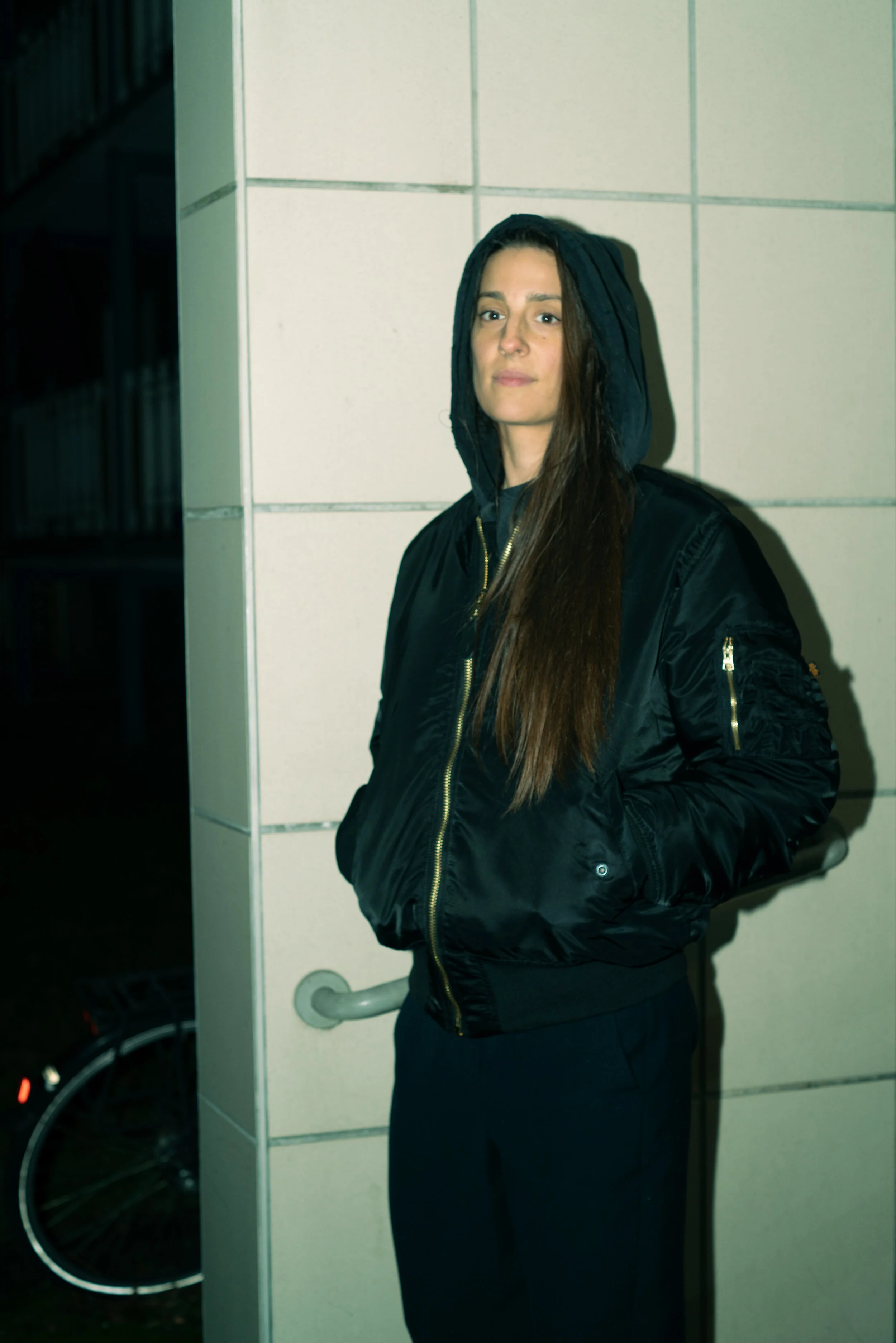 Woman with long brown hair wearing black hoodie and bomber jacket standing against a white tiled wall at night.