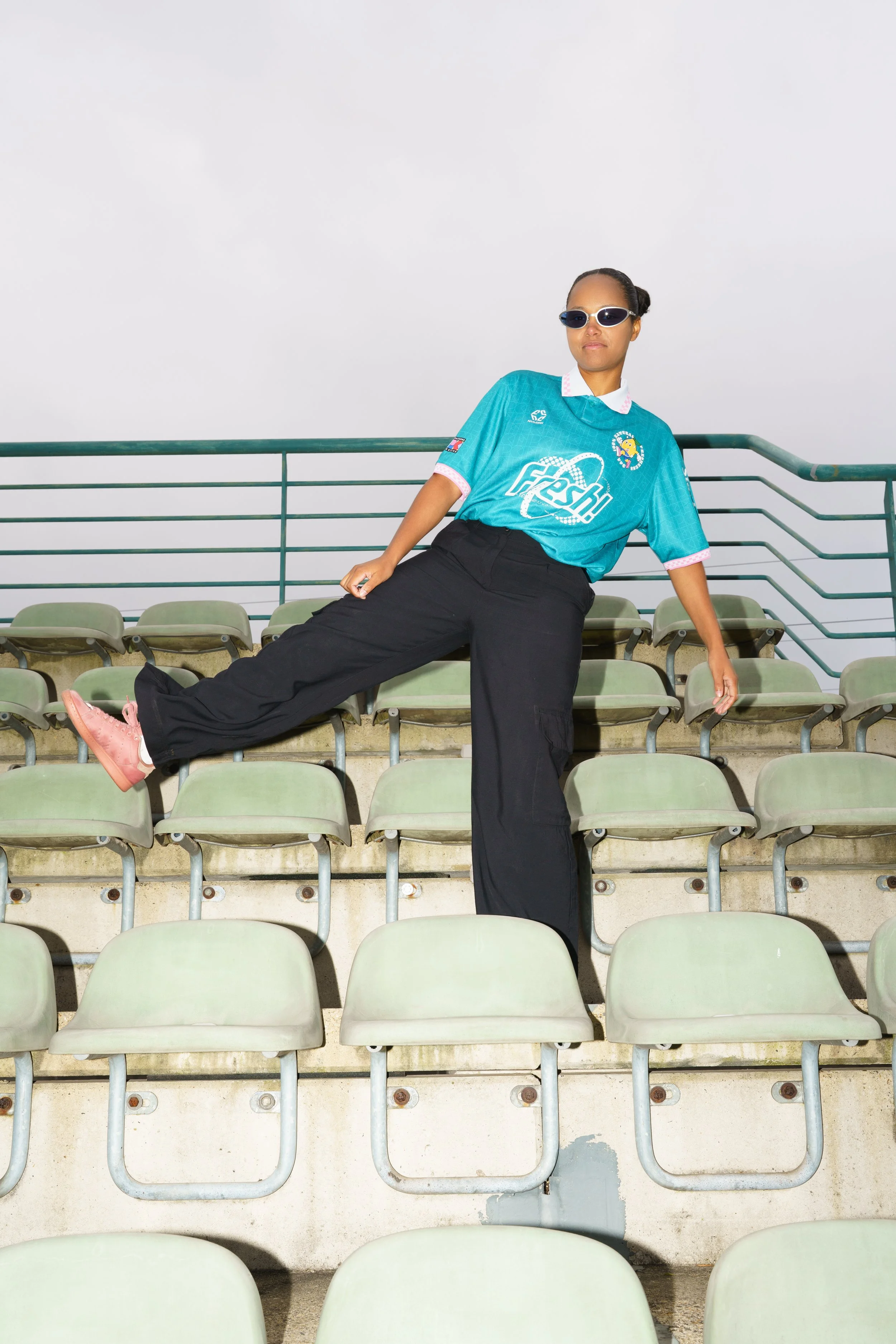 A woman wearing a turquoise sports jersey, black pants, pink sneakers, and sunglasses is posing on the empty seats of a stadium bleacher with a cloudy sky in the background.