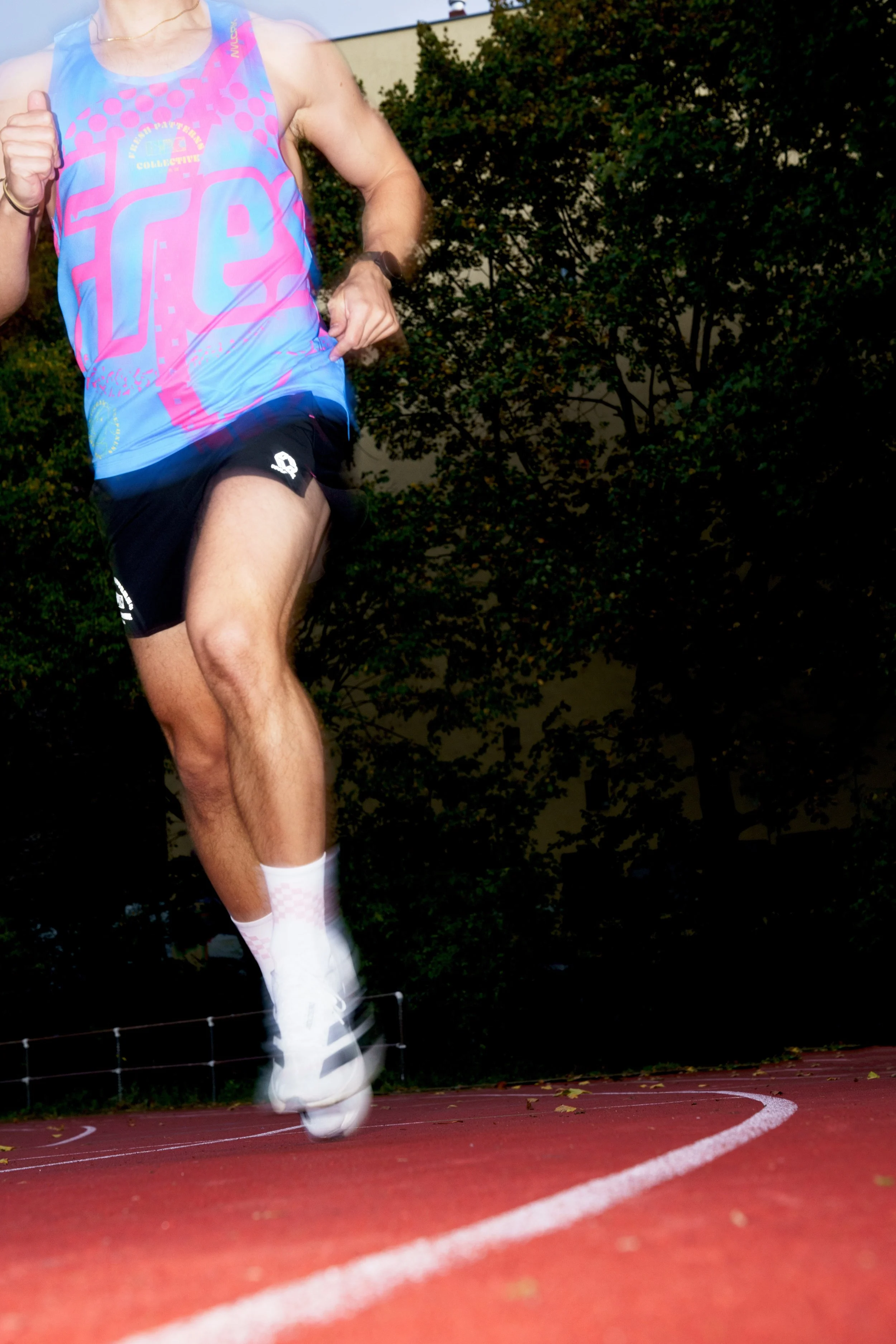 Close-up of a male runner in mid-air during a race on a track. He is wearing a colorful, sleeveless athletic shirt, black shorts, white running shoes, and white ankle socks. The background shows trees and a building.