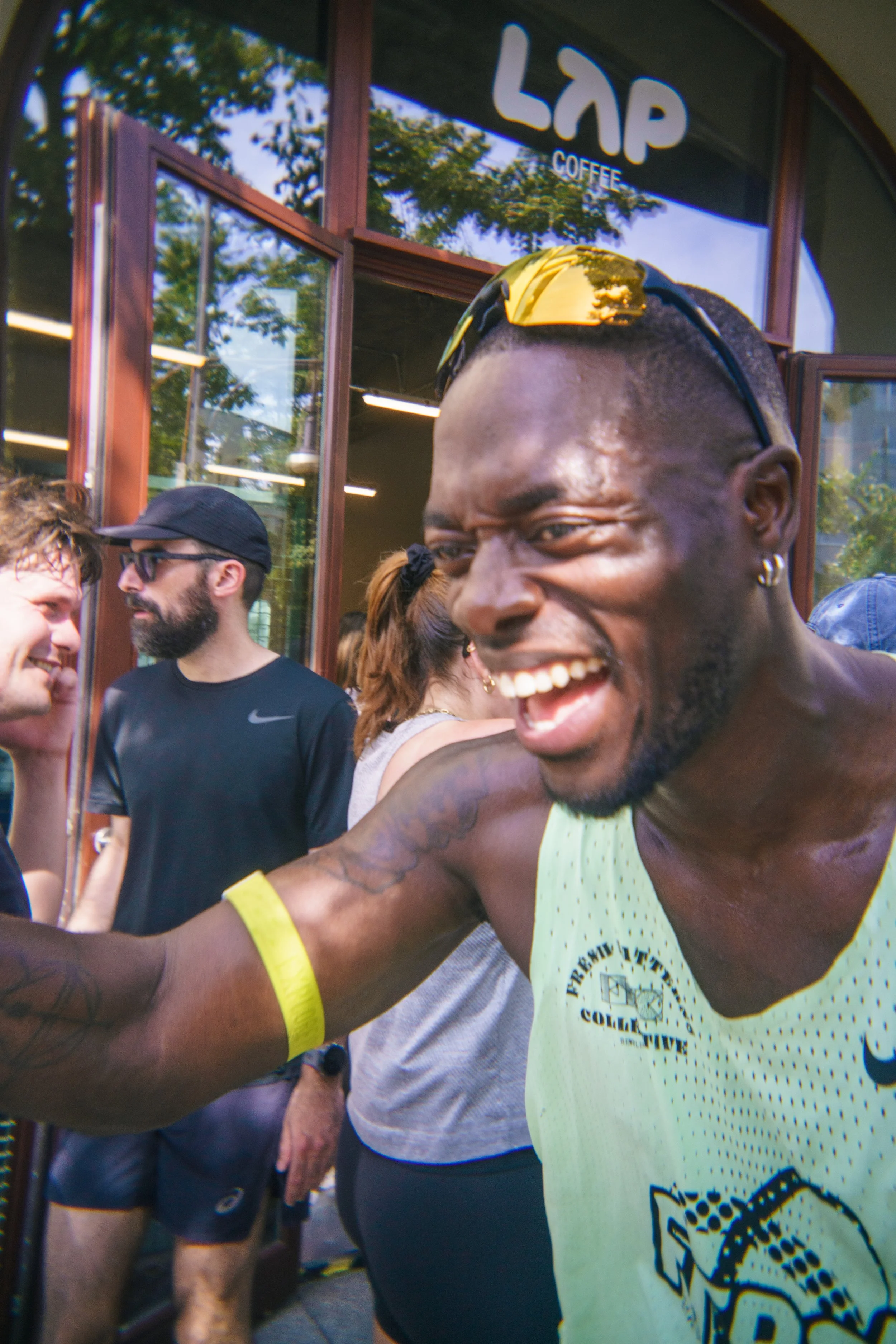 A group of people, including a smiling man with sunglasses on his head, outside a coffee shop. The man in the foreground is wearing a yellow athletic tank top and appears to be celebrating or happy, with others in the background engaging in conversat