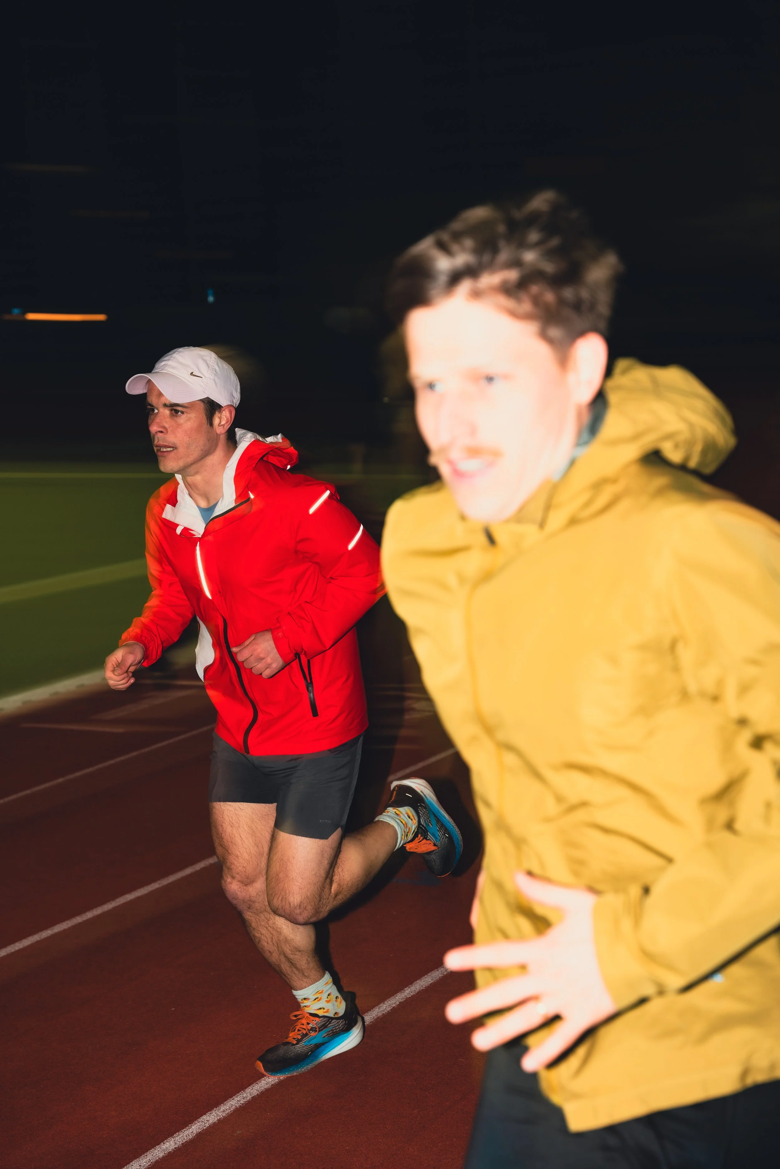 Two men running on a track at night, one in a red jacket and white cap, the other in a yellow jacket.