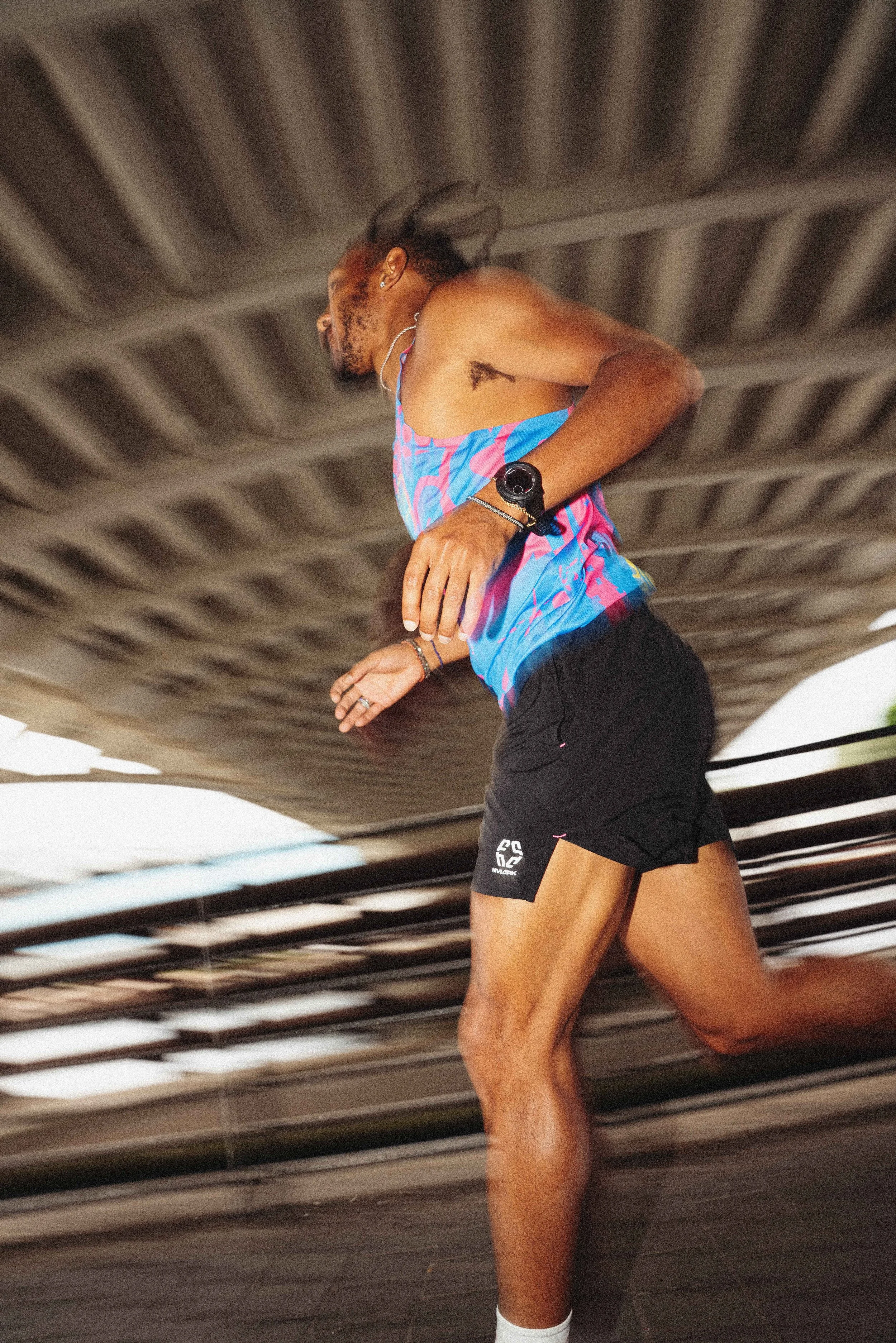 A male runner with a muscular build, wearing a colorful tank top, black shorts, white socks, and sports watch, running indoors.