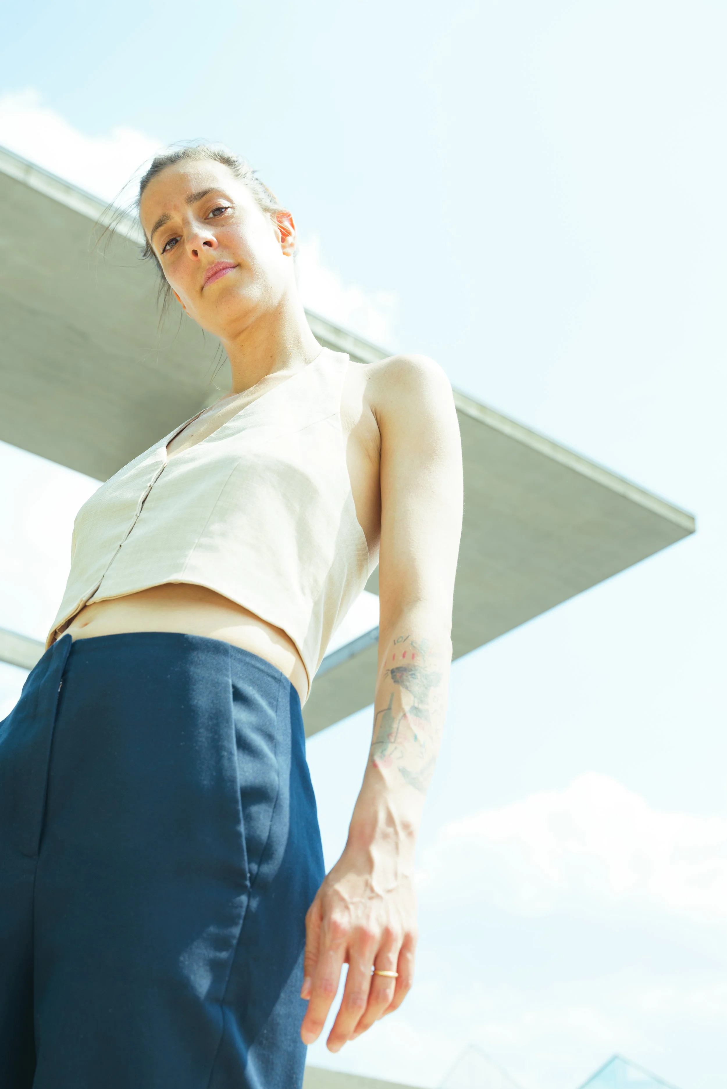 Low angle view of a young woman with a tattooed arm, wearing a sleeveless beige top and navy pants, standing outdoors under a concrete structure on a bright day.