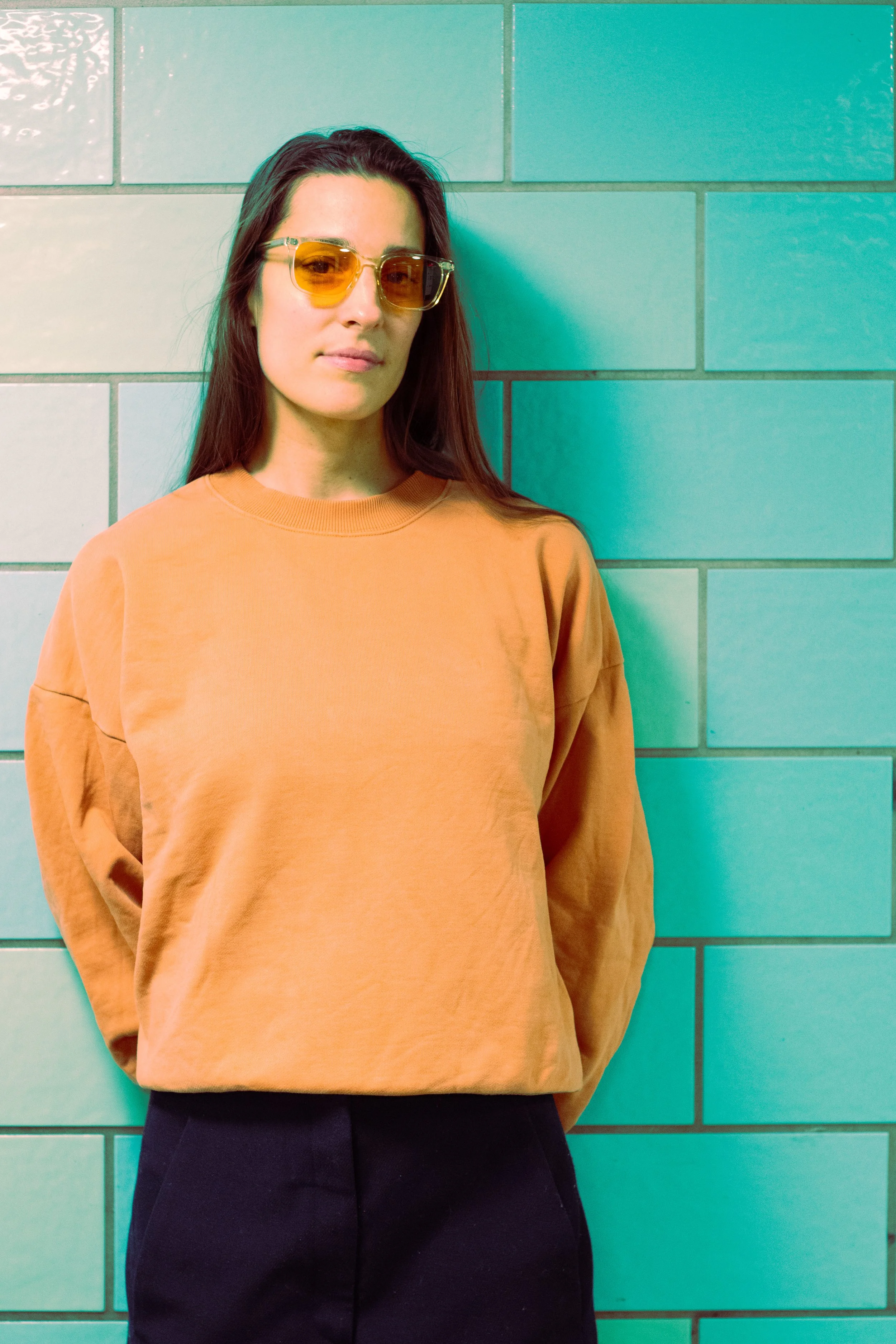 A woman with long dark hair wearing yellow-tinted sunglasses, an orange sweatshirt, and black pants, standing against a teal tiled wall.