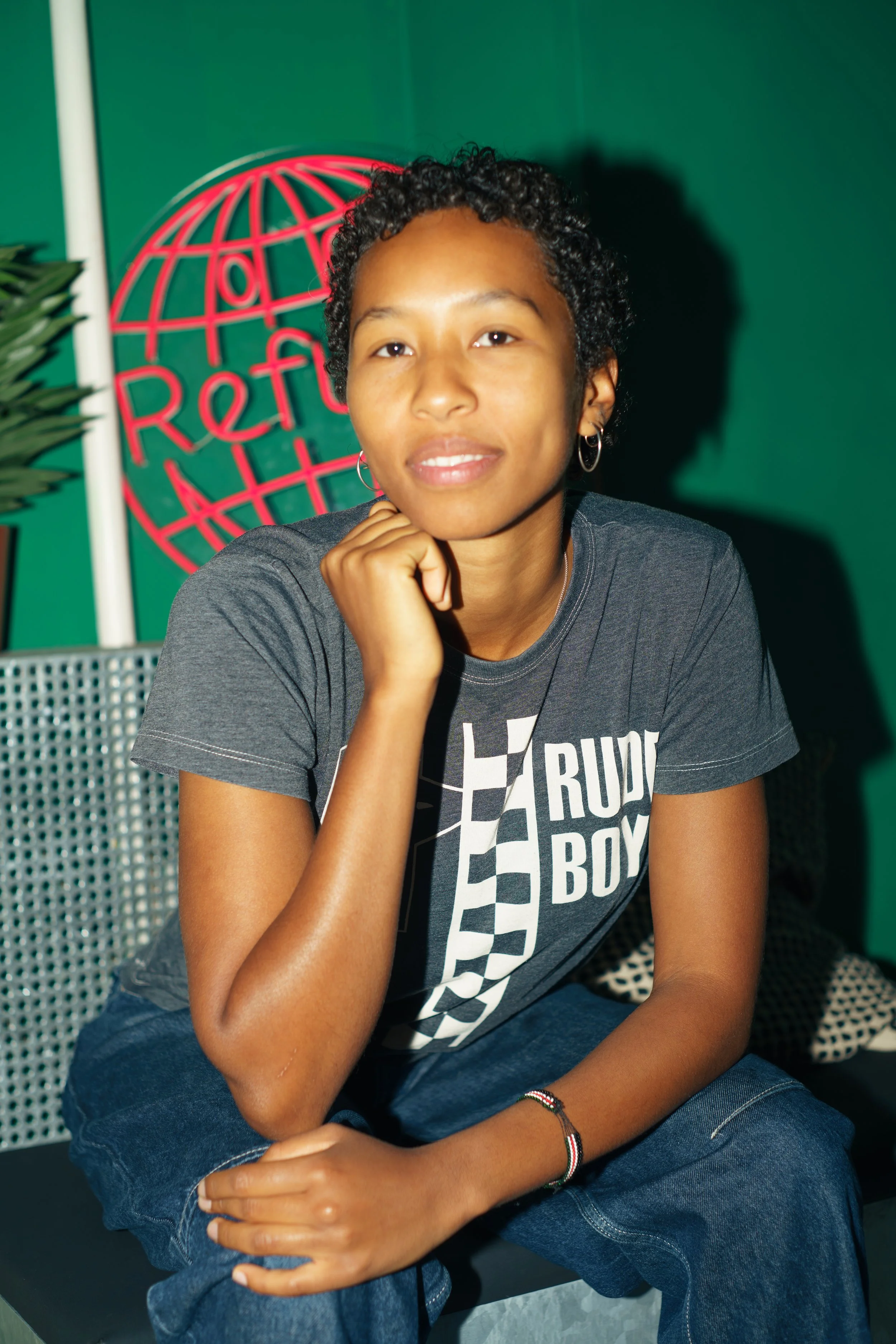Young woman with short curly hair and hoop earrings sitting indoors against a green background with a red globe logo that says 'Refugee' behind her, wearing a dark gray T-shirt and jeans, resting her chin on her hand.