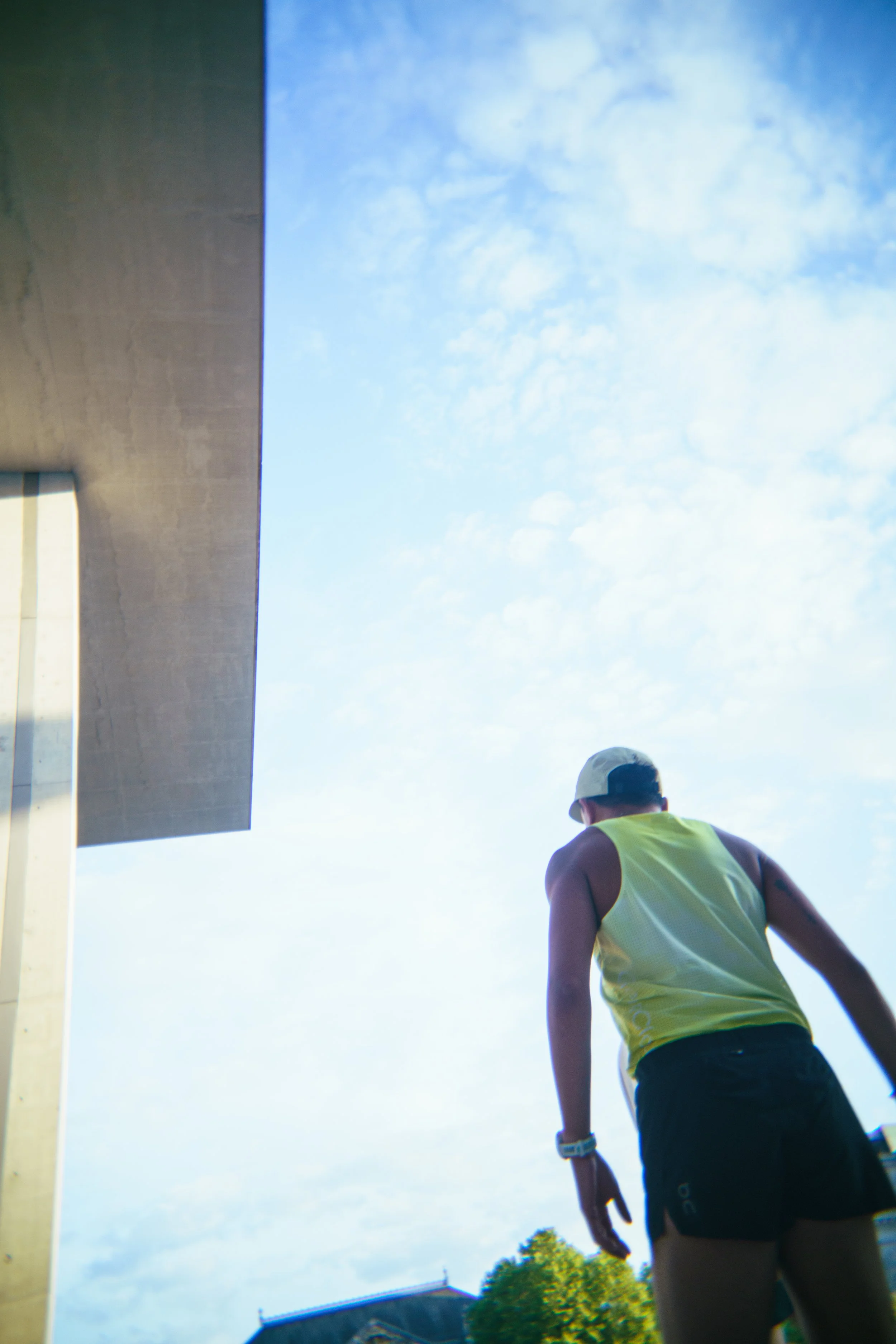 A person in athletic clothing, wearing a yellow tank top, black shorts, a white cap, and a watch, is seen from behind outdoors on a sunny day with a blue sky and scattered clouds, near a modern building with a concrete and glass facade.