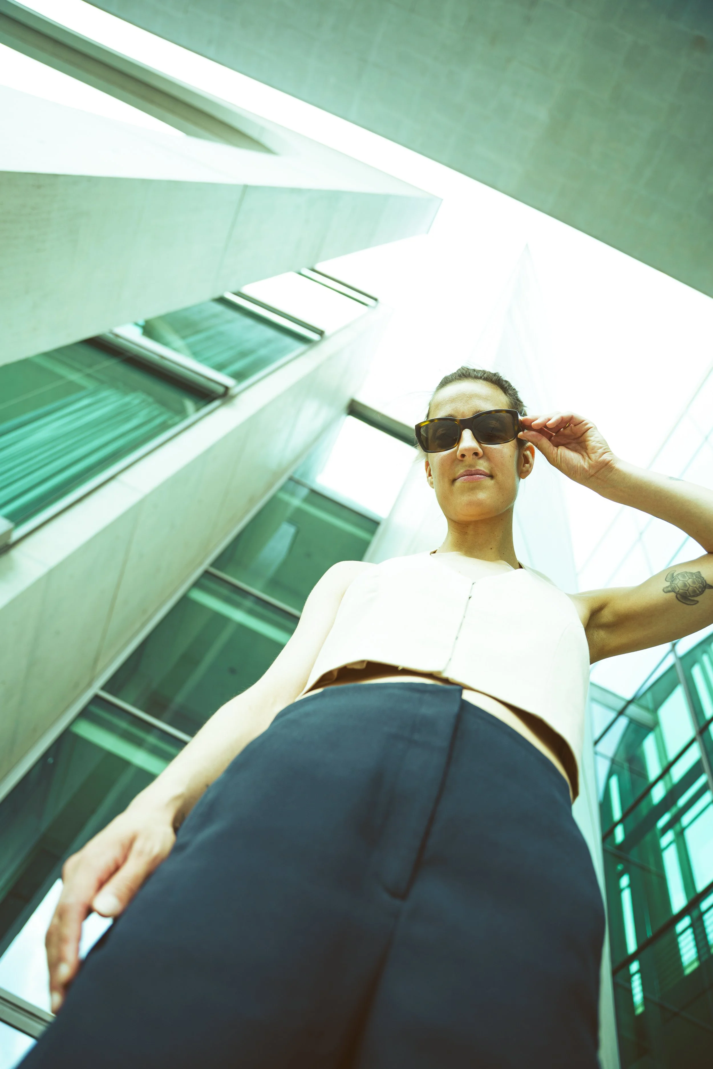 A woman standing outdoors between modern glass buildings, wearing sunglasses, a sleeveless top, and a dark skirt, seen from a low-angle shot.