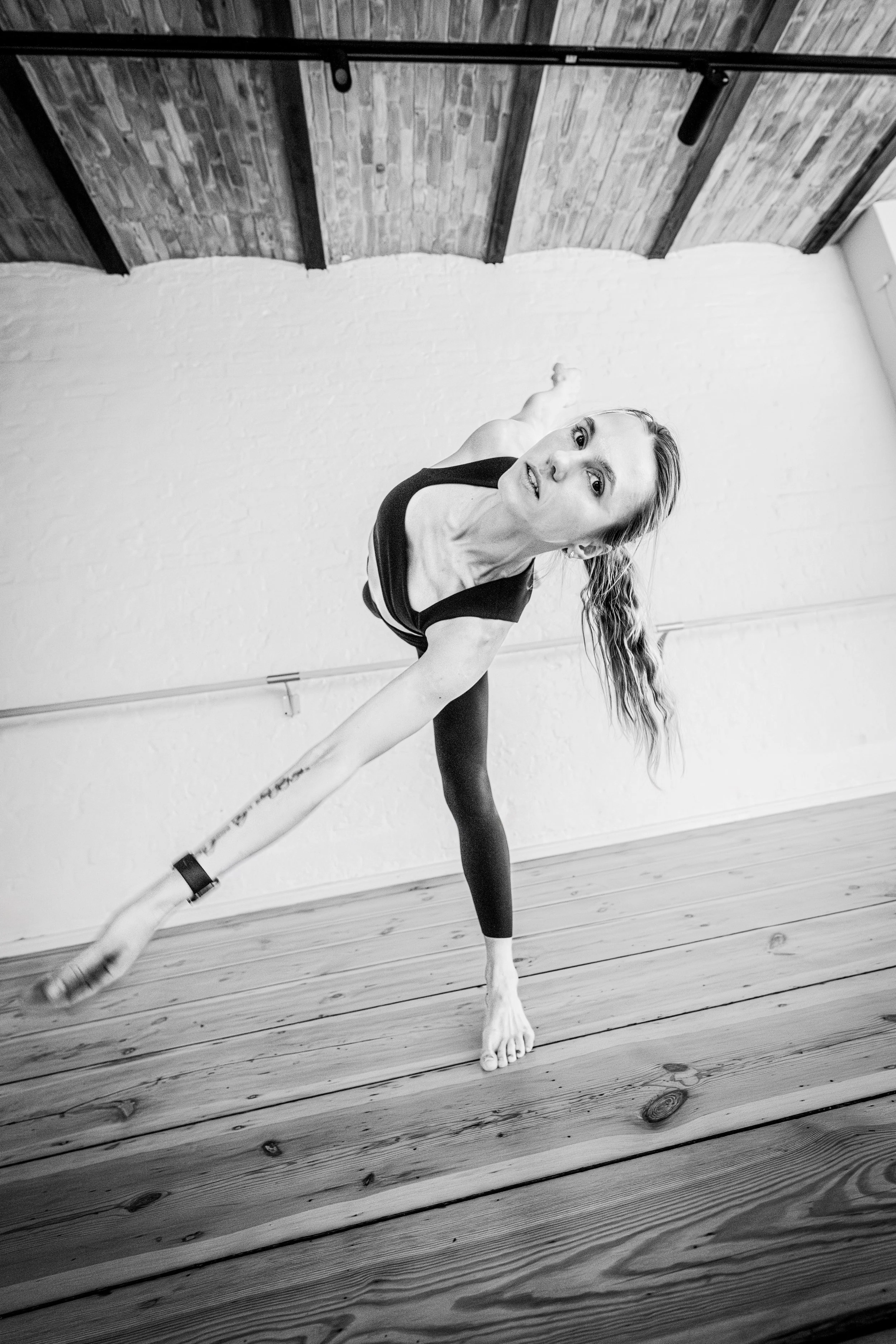 A woman practicing yoga in a studio with wooden floors, a white brick wall, and a wooden ceiling, performing a side stretch pose.