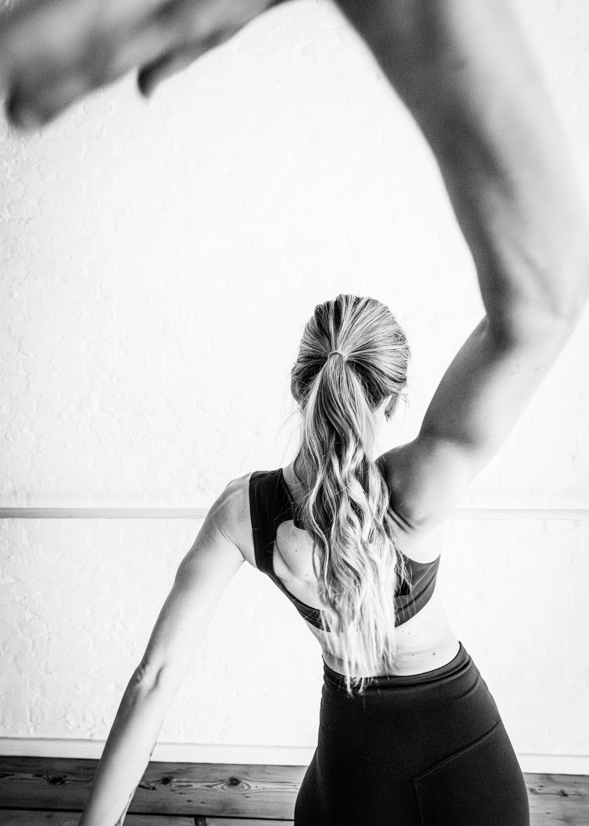 Black and white photo of a woman with long wavy hair tied back, practicing yoga or stretching against a plain wall, with her arm raised above her head.