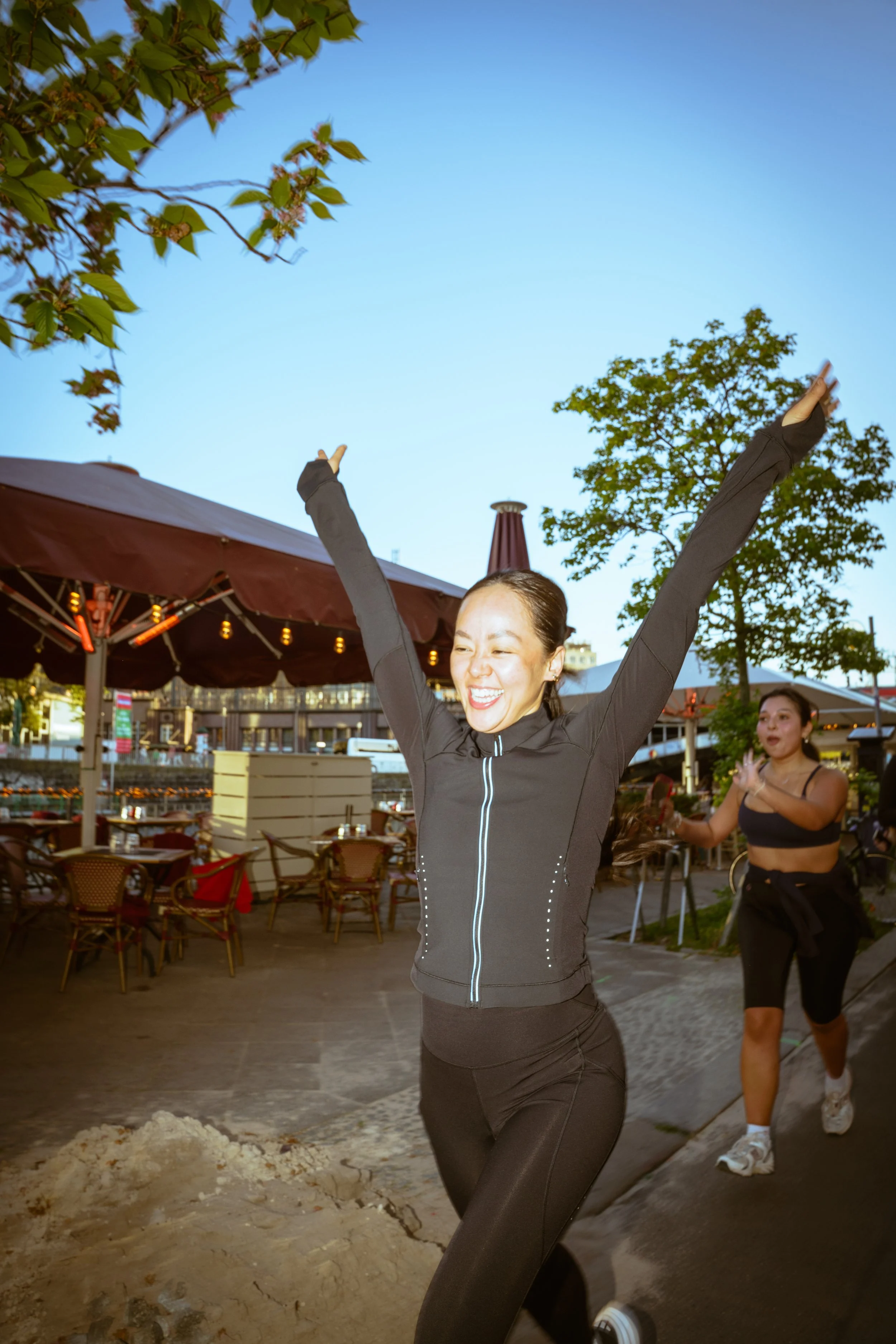 A woman with arms raised celebrating outdoors in the evening, near a patio with tables and umbrellas, another woman jogging behind her, trees, and a clear sky.