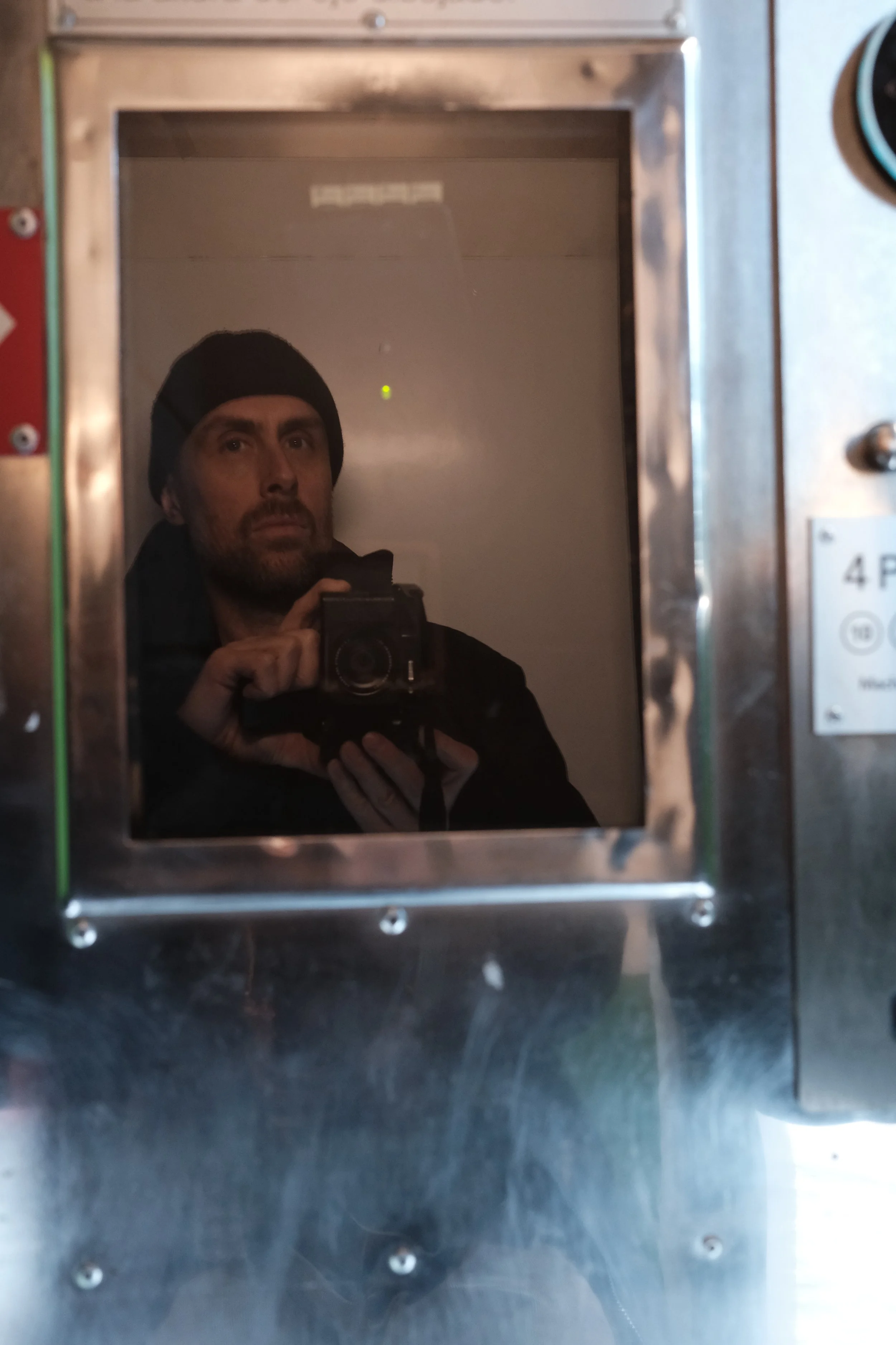 A person taking a photo of their reflection in a small, rectangular mirror on a stainless steel wall, wearing a black beanie and dark clothing.