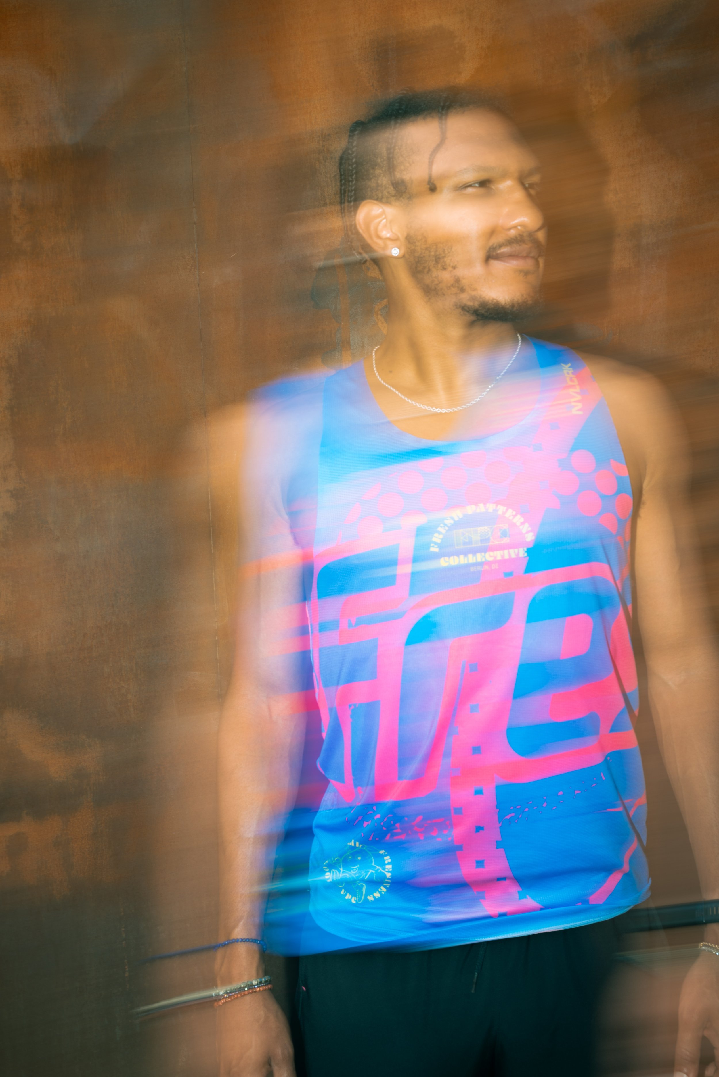 A person with braided hair, wearing a colorful sleeveless shirt with pink and blue designs, standing in front of a wooden background. The person has earrings, a necklace, and bracelets, with a slight smile on their face. The photo has motion blur eff