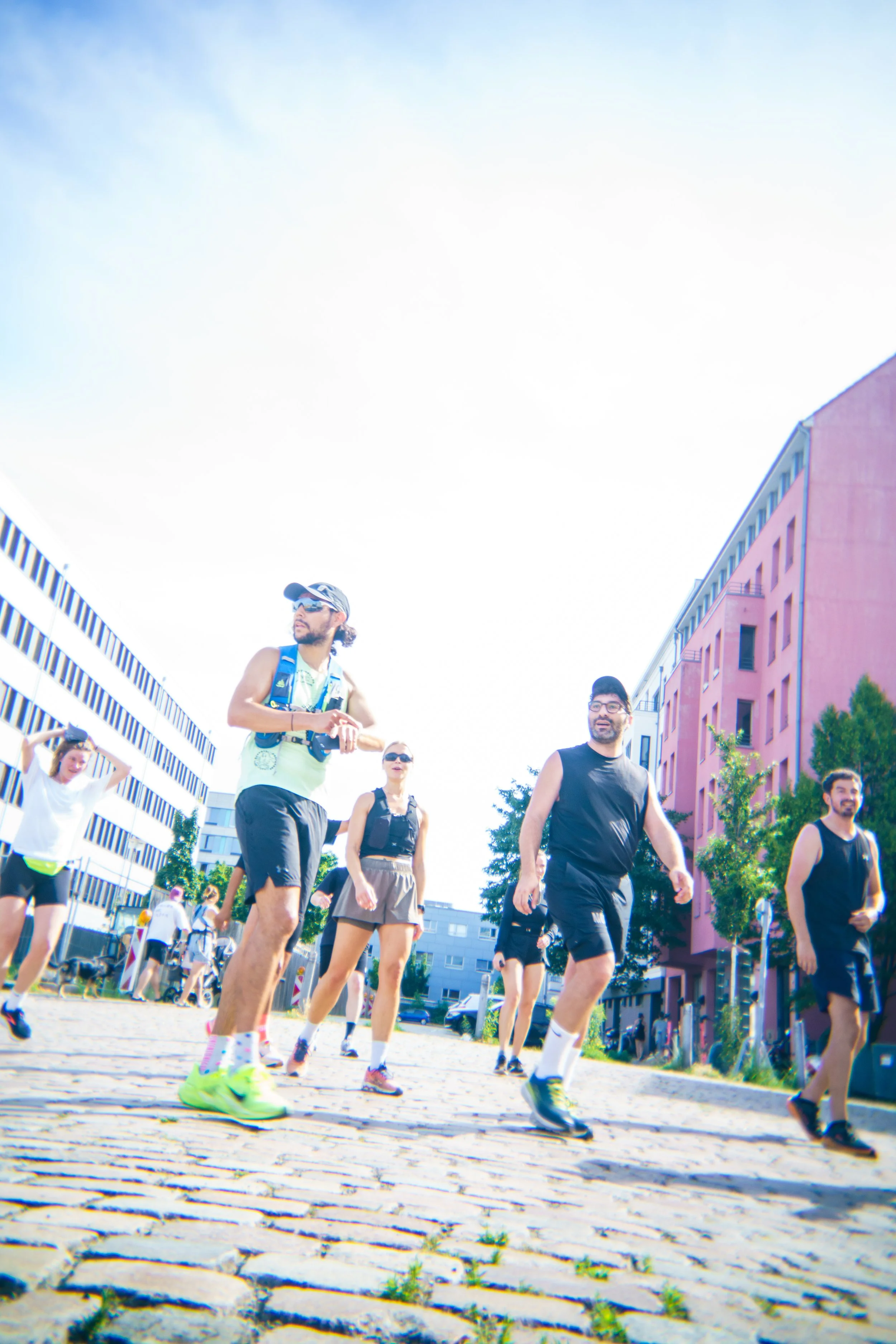 A group of people in athletic clothing walking and jogging outdoors on a sunny day in an urban area with colorful buildings and trees.