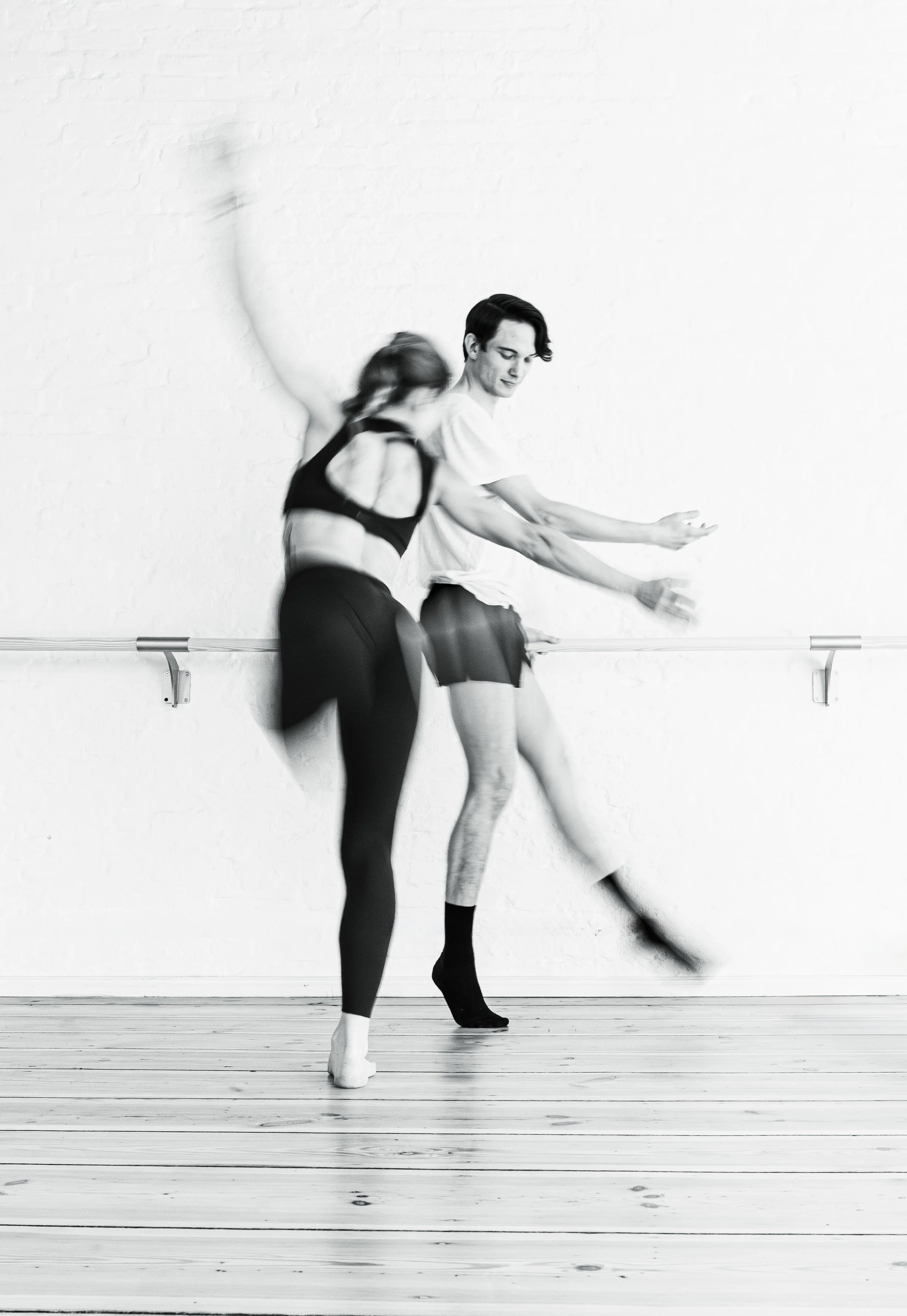 Two ballet dancers practicing at the barre in a dance studio, one woman and one man, captured in motion in black and white.