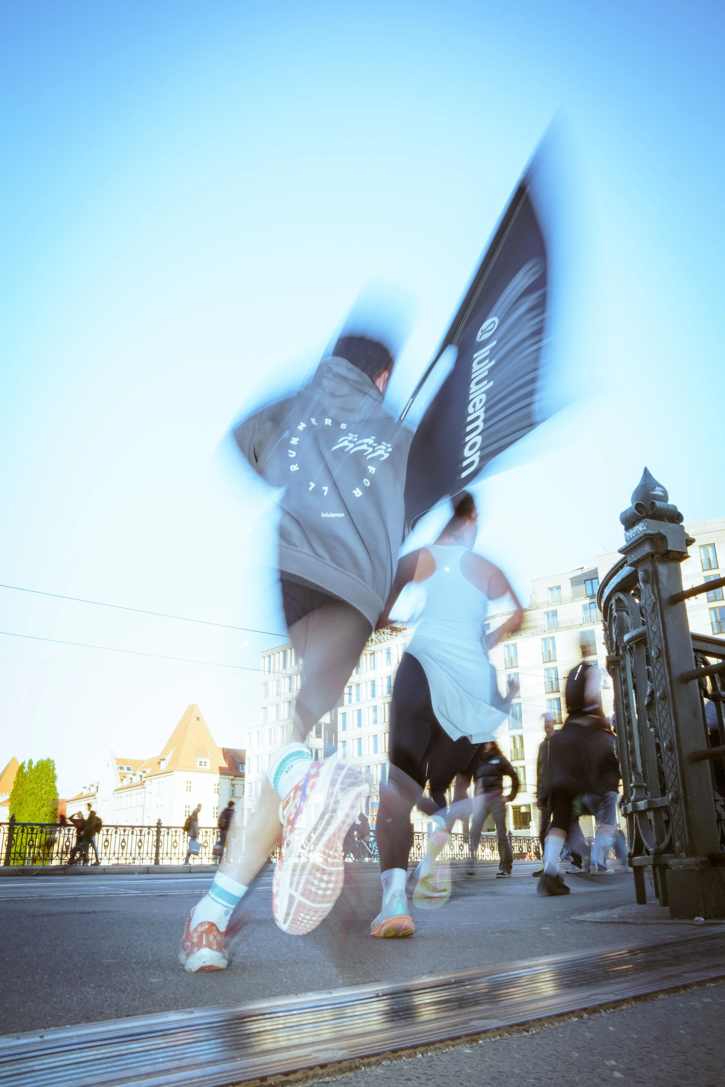 People running on a city street during a race, with some runners appearing blurred due to motion, and others visible in the background near buildings and a fence.