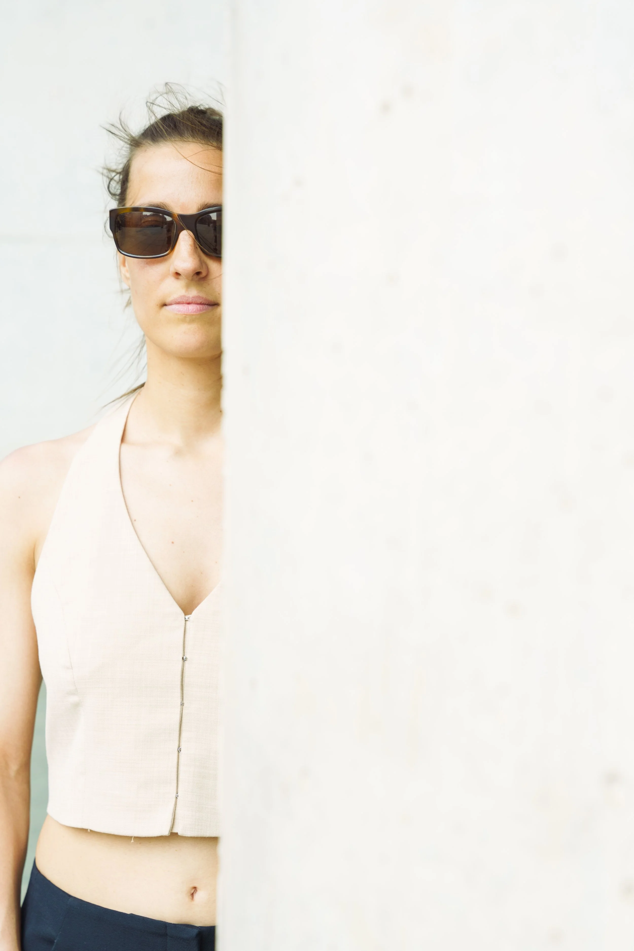 A woman with dark sunglasses and a sleeveless beige top is partially hidden behind a white wall.