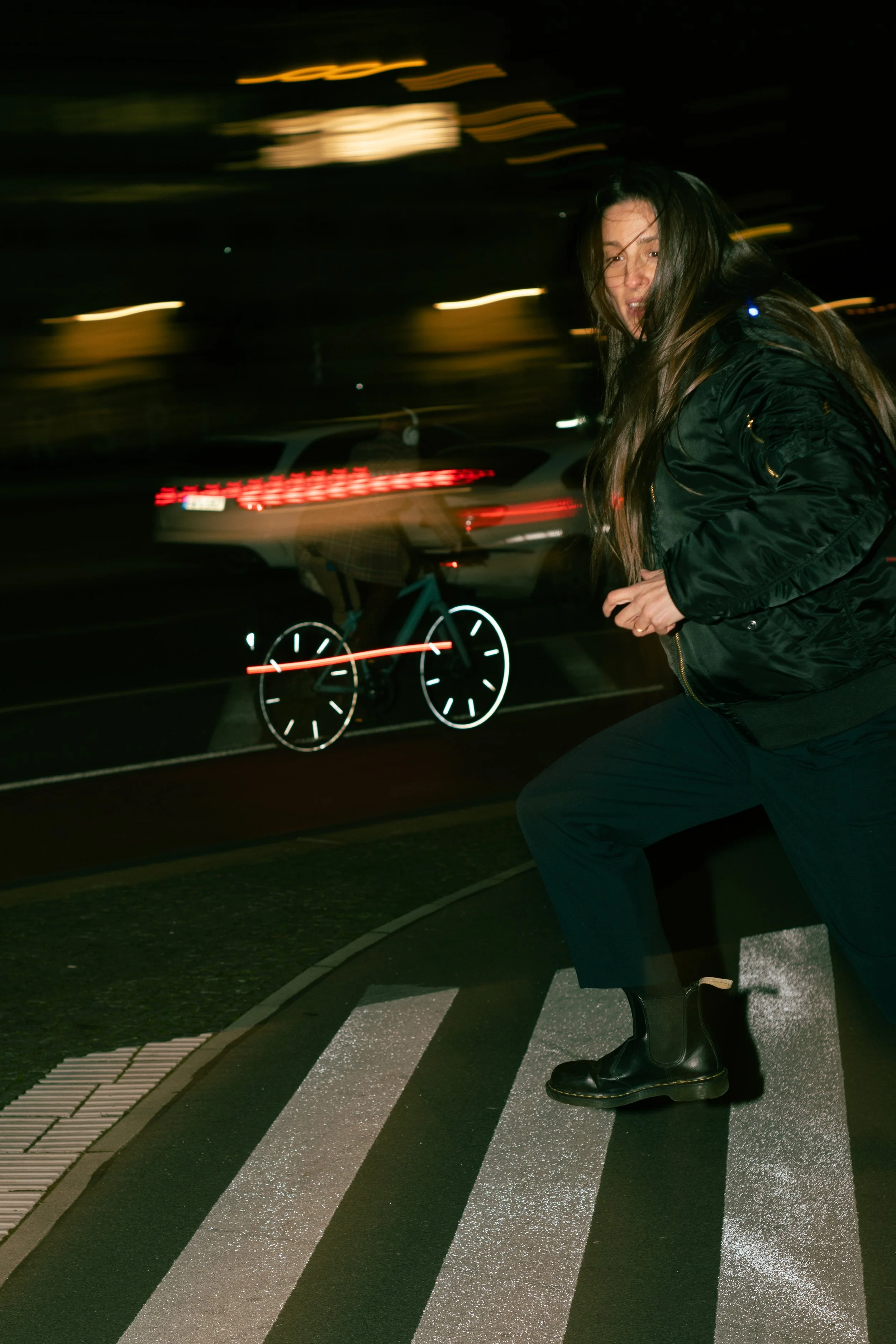 A woman wearing a black jacket and black boots crossing a pedestrian crosswalk at night, with a blurred car and a cyclist in the background.