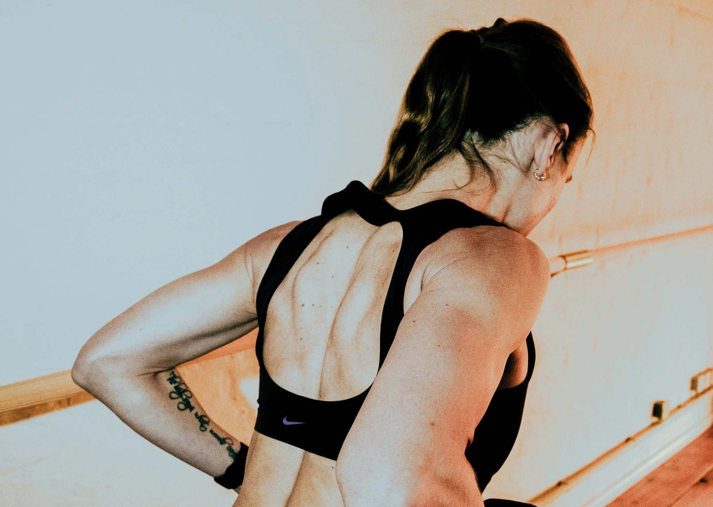 A woman with dark hair in a braid, wearing a black sports bra and Nike logo, is leaning forward, appearing to stretch or exercise indoors.