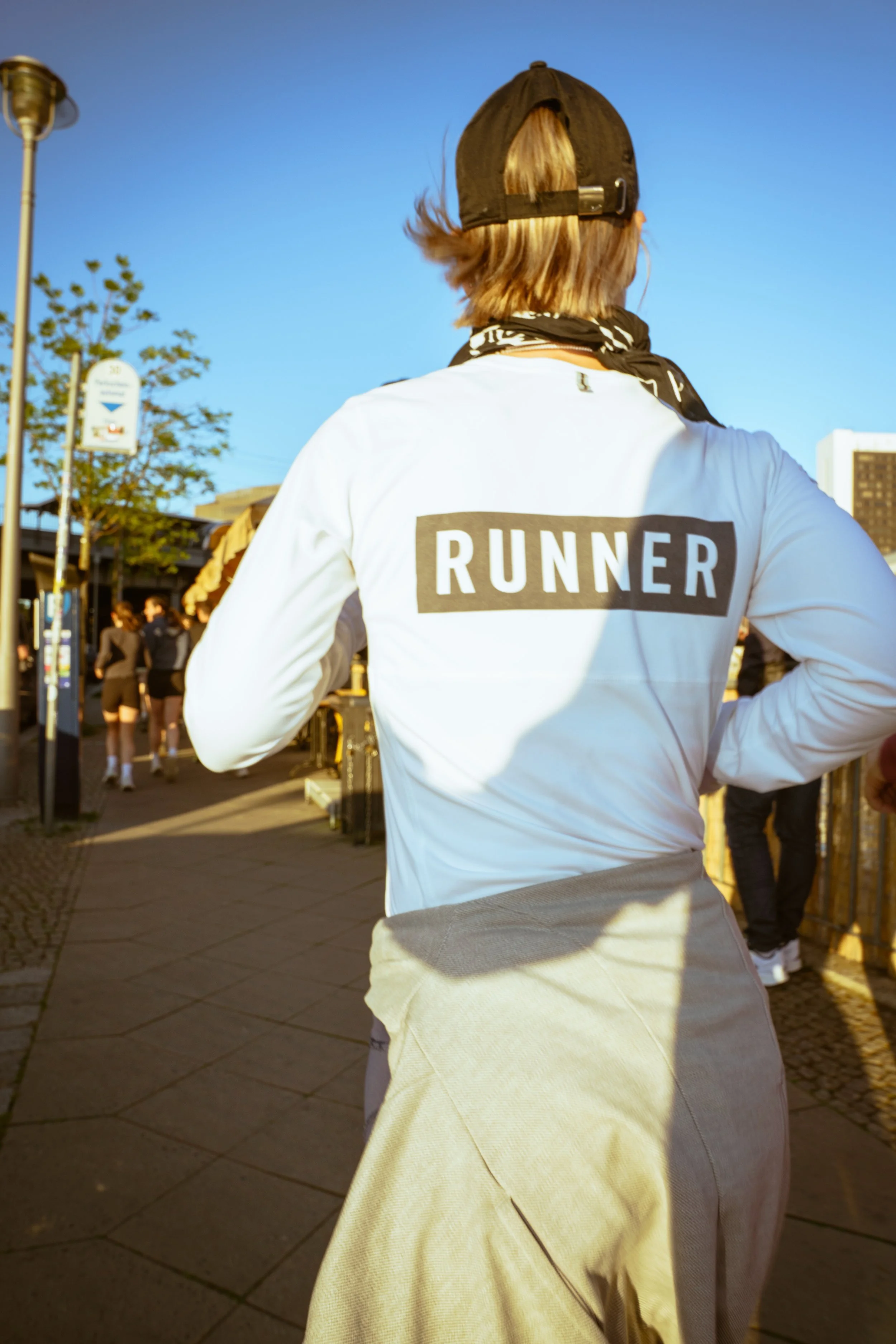 Back view of a runner in a white shirt with 'RUNNER' written on it, wearing a baseball cap and a black bandana around the neck, during a daytime outdoor run.