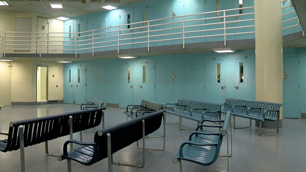 Empty waiting area with black and metal benches, light blue walls, and a second-floor walkway with white railing in a correctional facility or detention center.