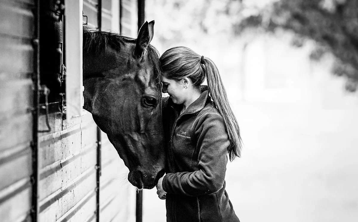 A woman with long hair tied in a ponytail leaning her forehead against a horse's forehead outside a wooden stable.