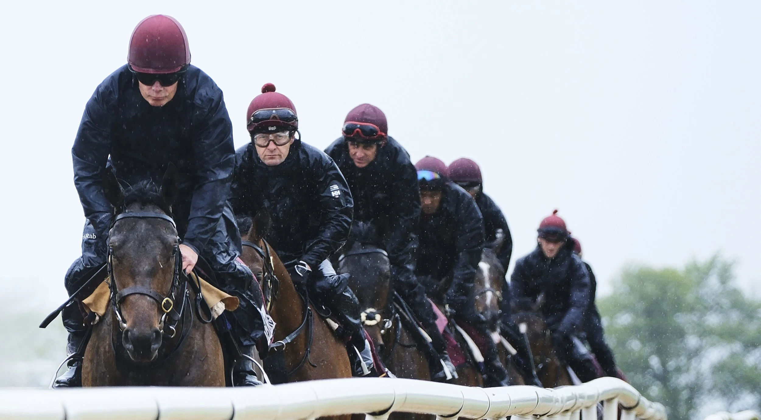 Racehorses with jockeys riding along a racetrack during a rainy day.