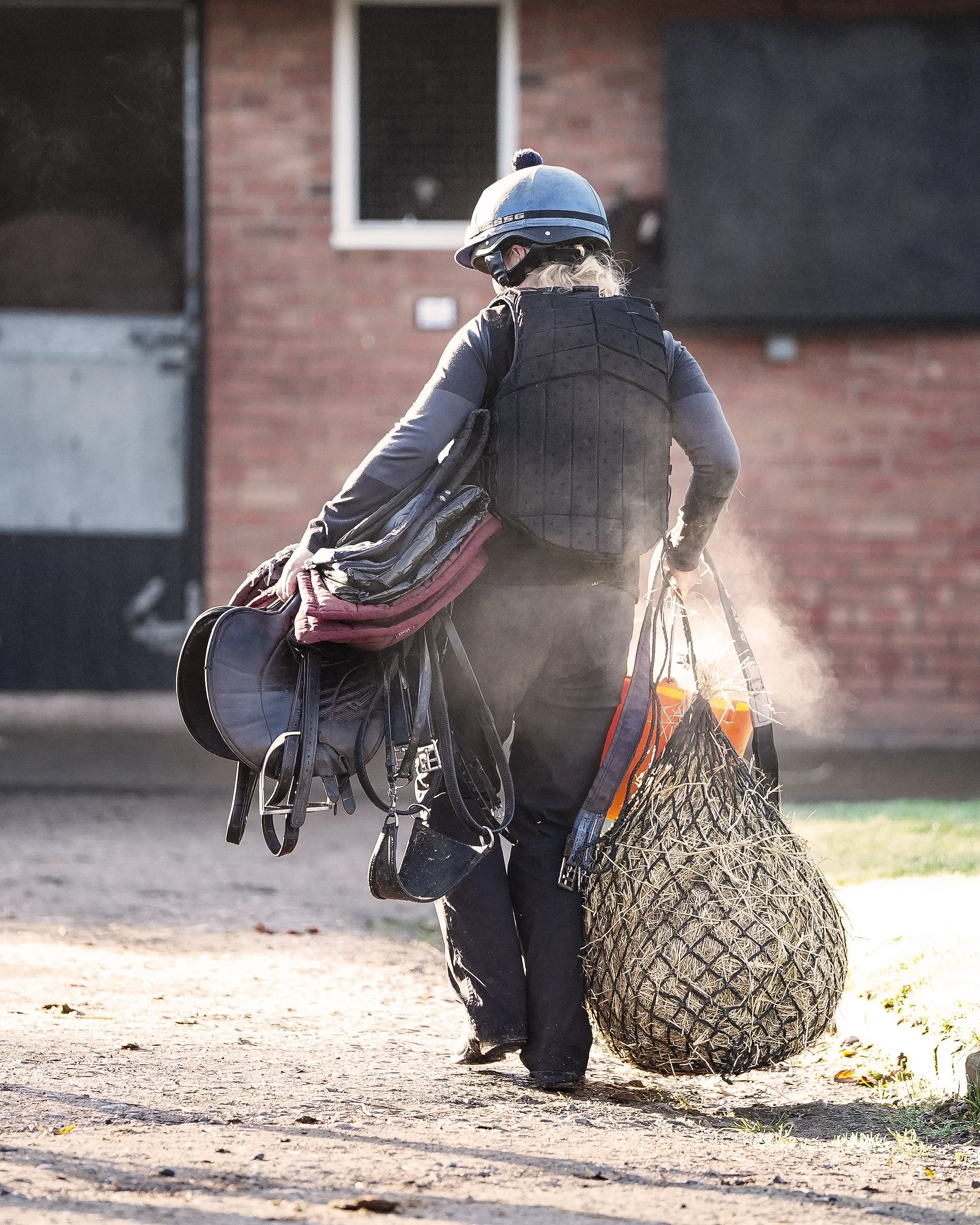 A person walking away from the camera, carrying horse riding gear and a large bag, wearing a helmet and a black vest, with a smoke or steam rising behind them on a dirt path in front of a brick building.