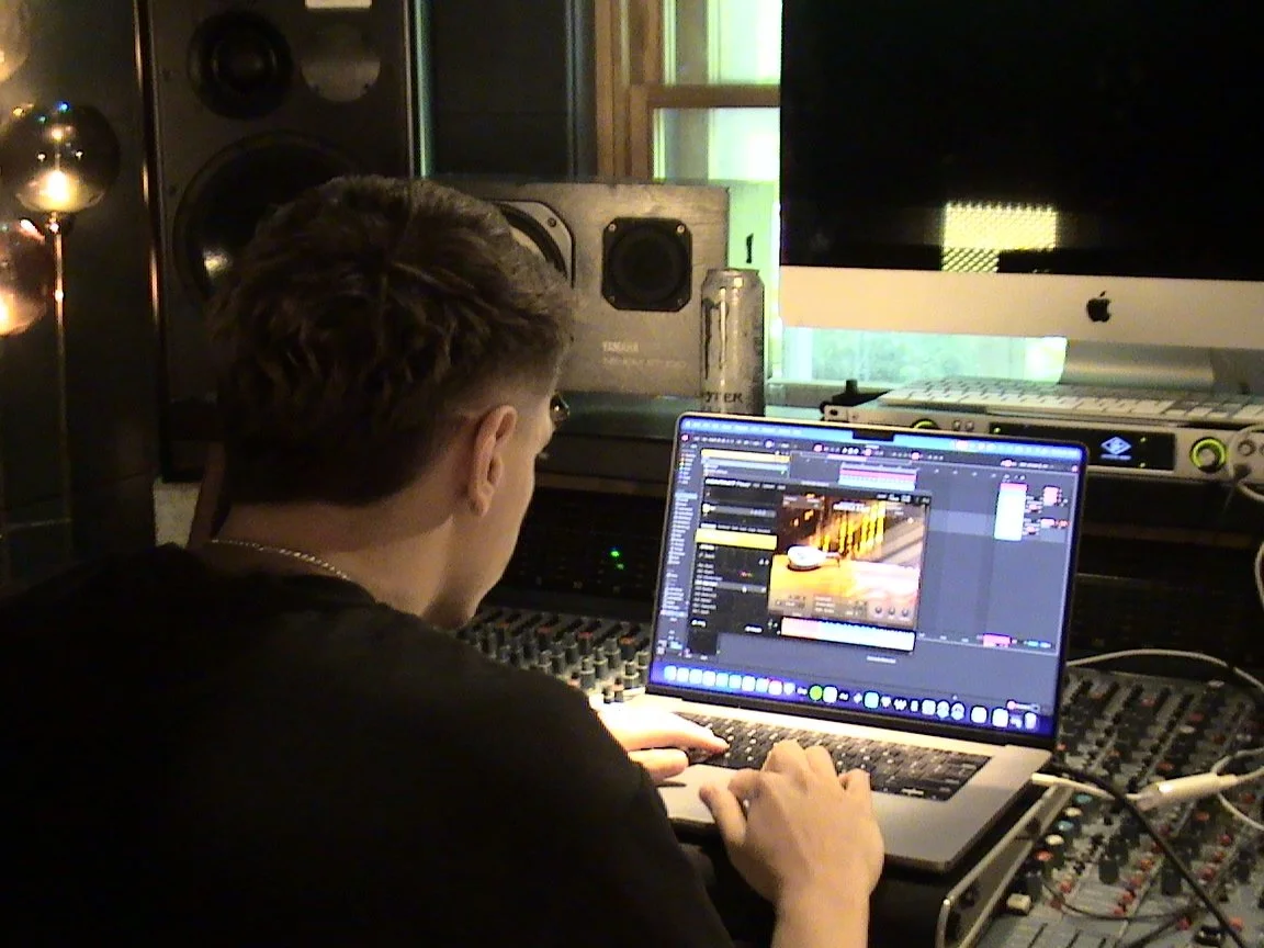 Person working on a laptop in a music studio, surrounded by speakers and audio equipment.