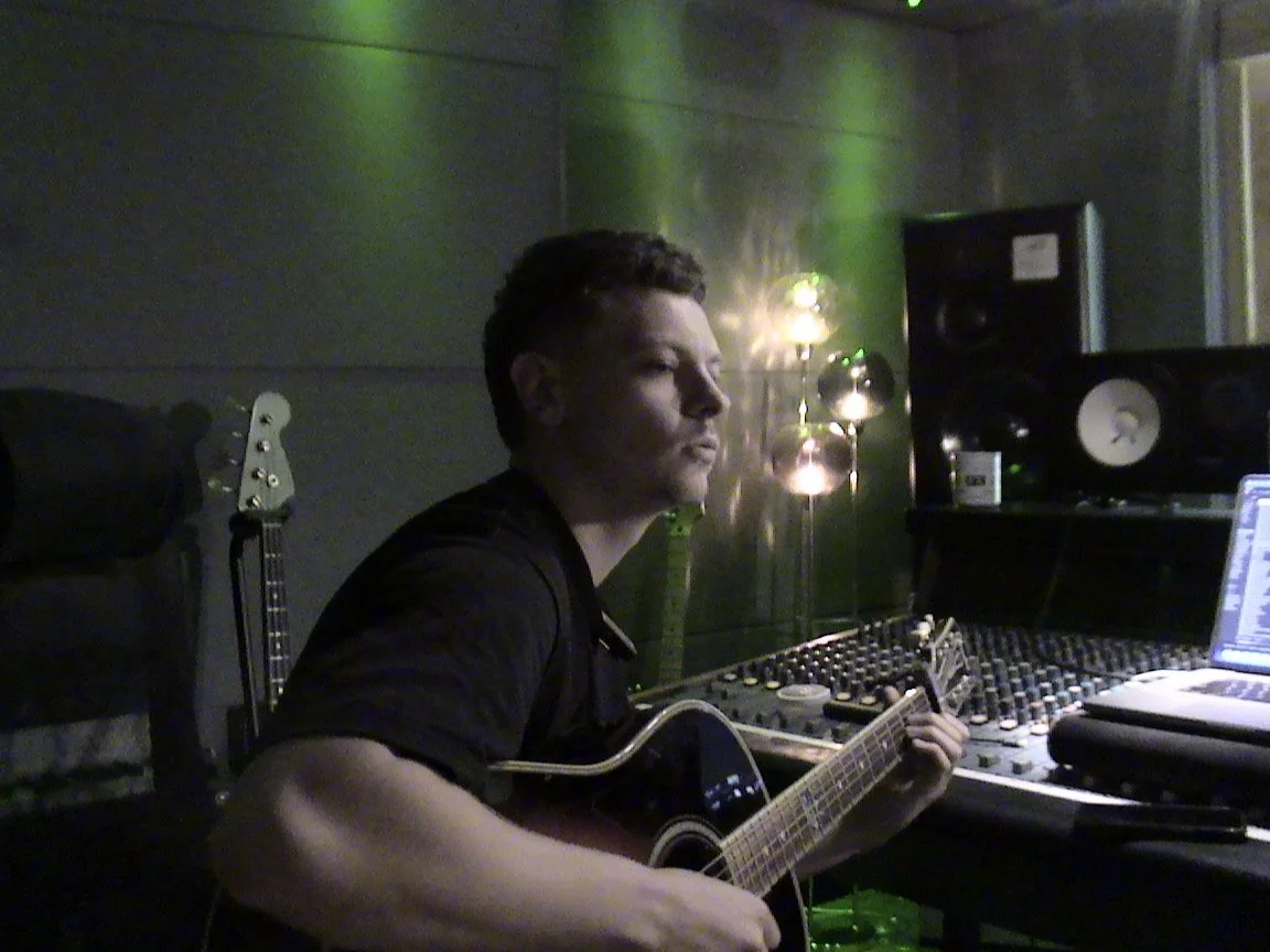 A man playing an acoustic guitar in a recording studio, with mixing equipment and studio speakers around him.