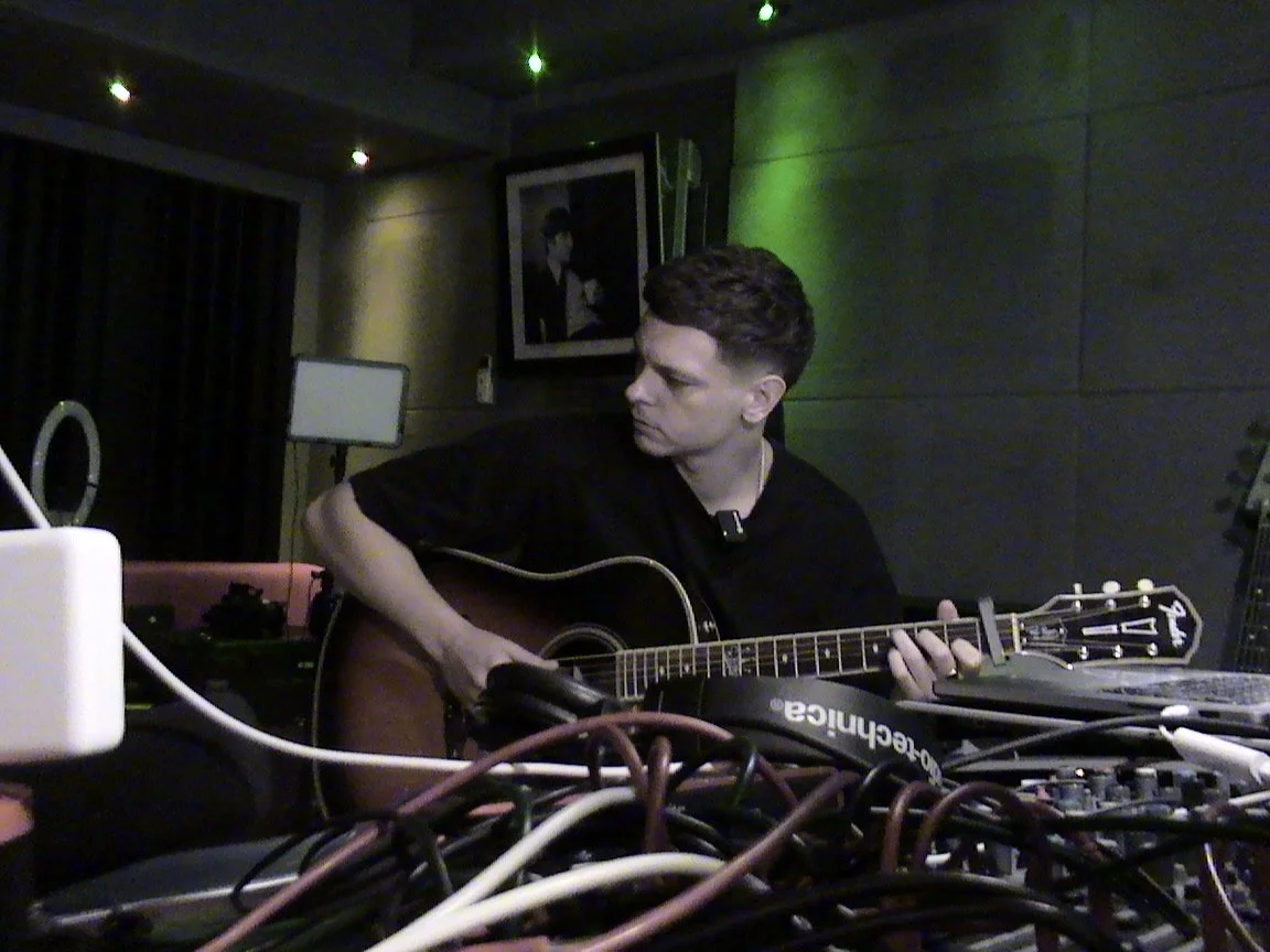 A man playing an acoustic guitar in a dimly lit room with electronic equipment and a picture of a man in sunglasses on the wall.