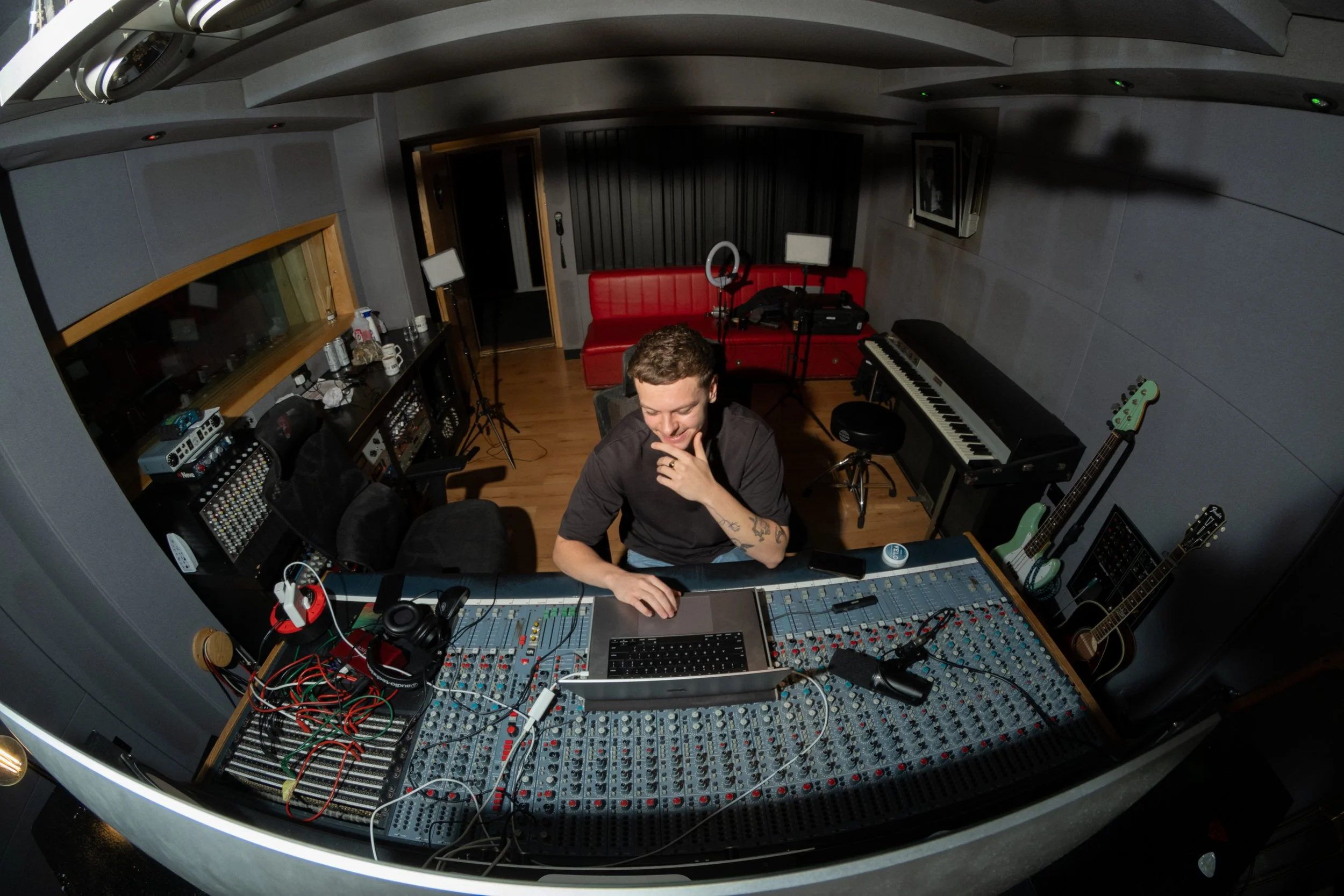 A young man in a recording studio, smiling and looking down at a mixing console. The studio has various musical instruments, equipment, and sound gear, including guitars, a keyboard, studio monitors, and microphones.