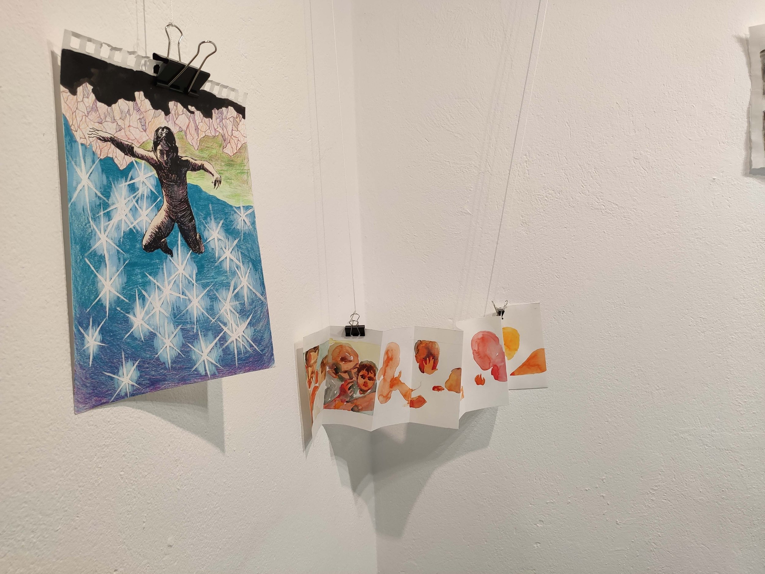 Colorful artwork displayed on a white wall, including a poster of a man in mid-air with arms outstretched, and a string of small watercolor paintings of abstract faces and shapes hanging from a wire.